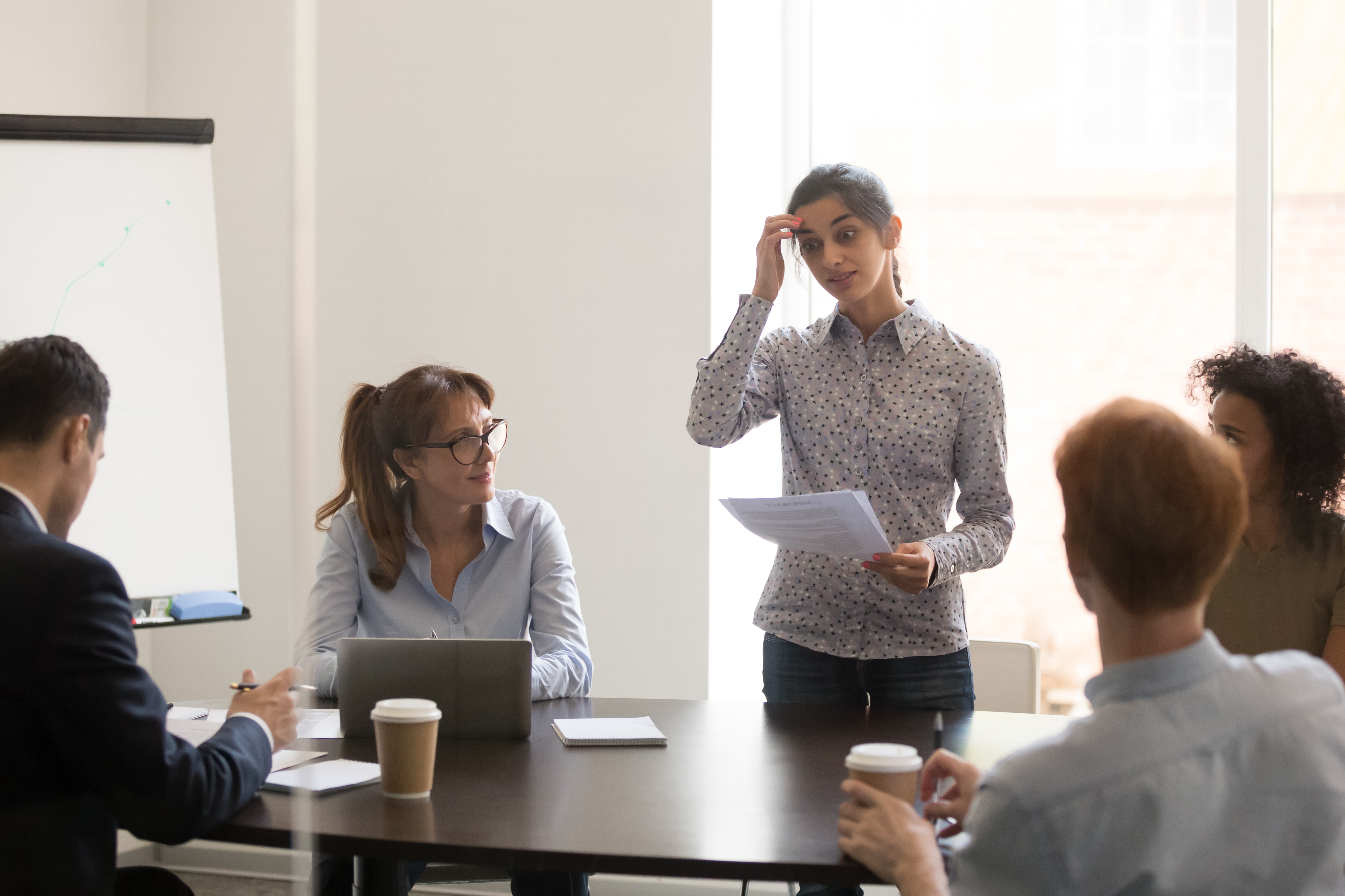 A woman stands holding papers, appearing to present to a group of seated colleagues in a meeting room