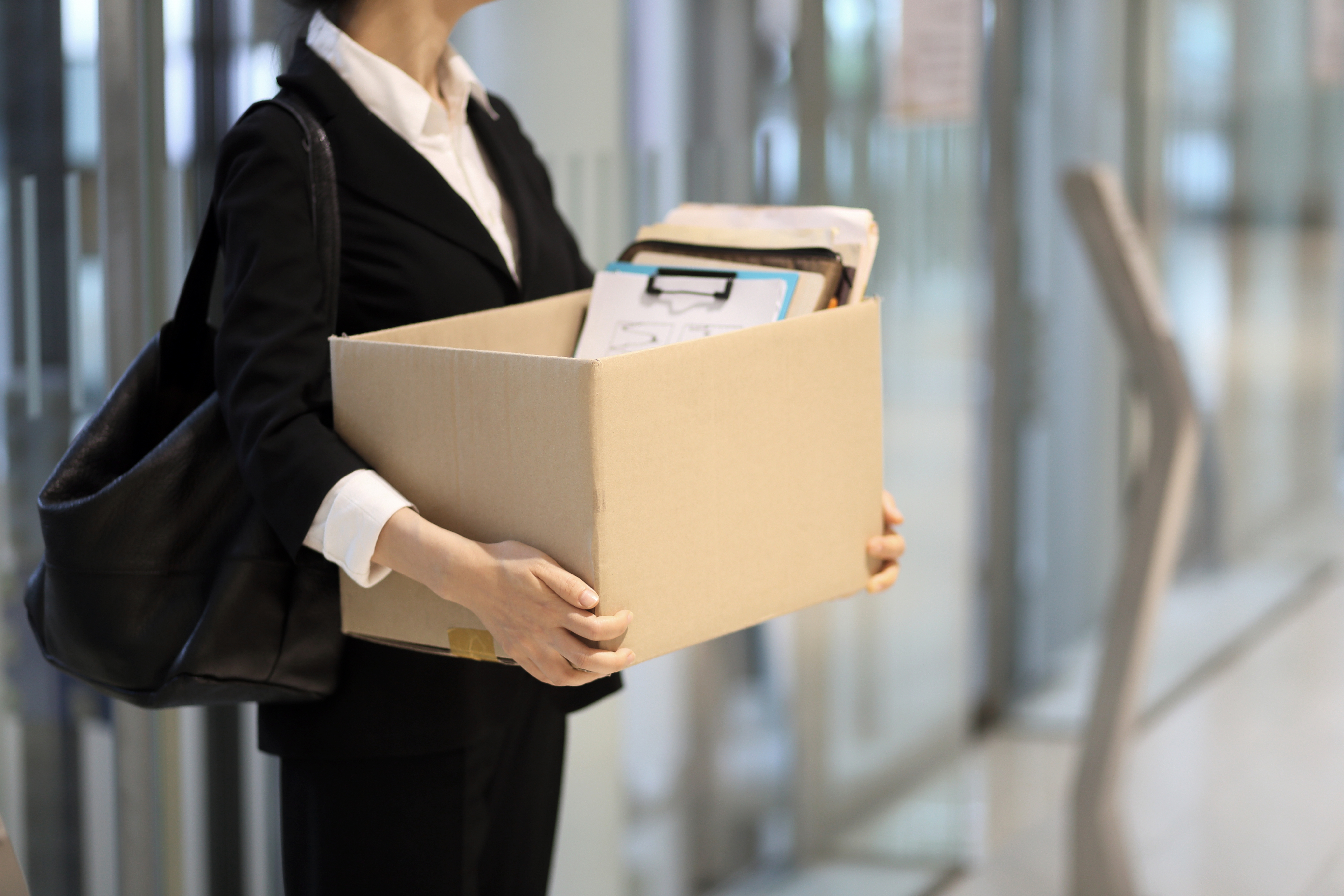 Person in business attire carrying a box filled with files and a clipboard in an office setting