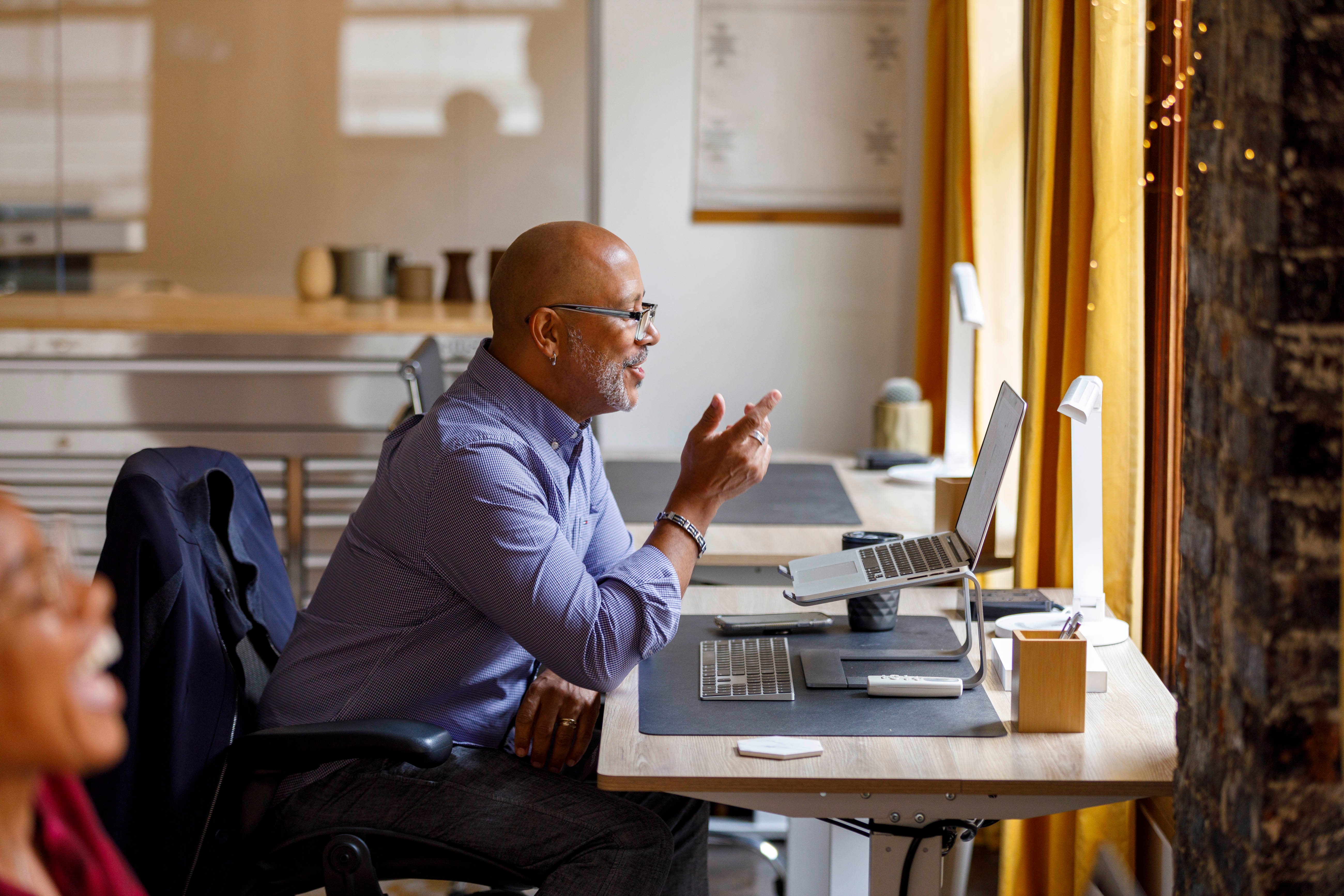 A man sits at a desk, smiling and speaking during a video call. Two monitors are on the desk, and a window is in front of him