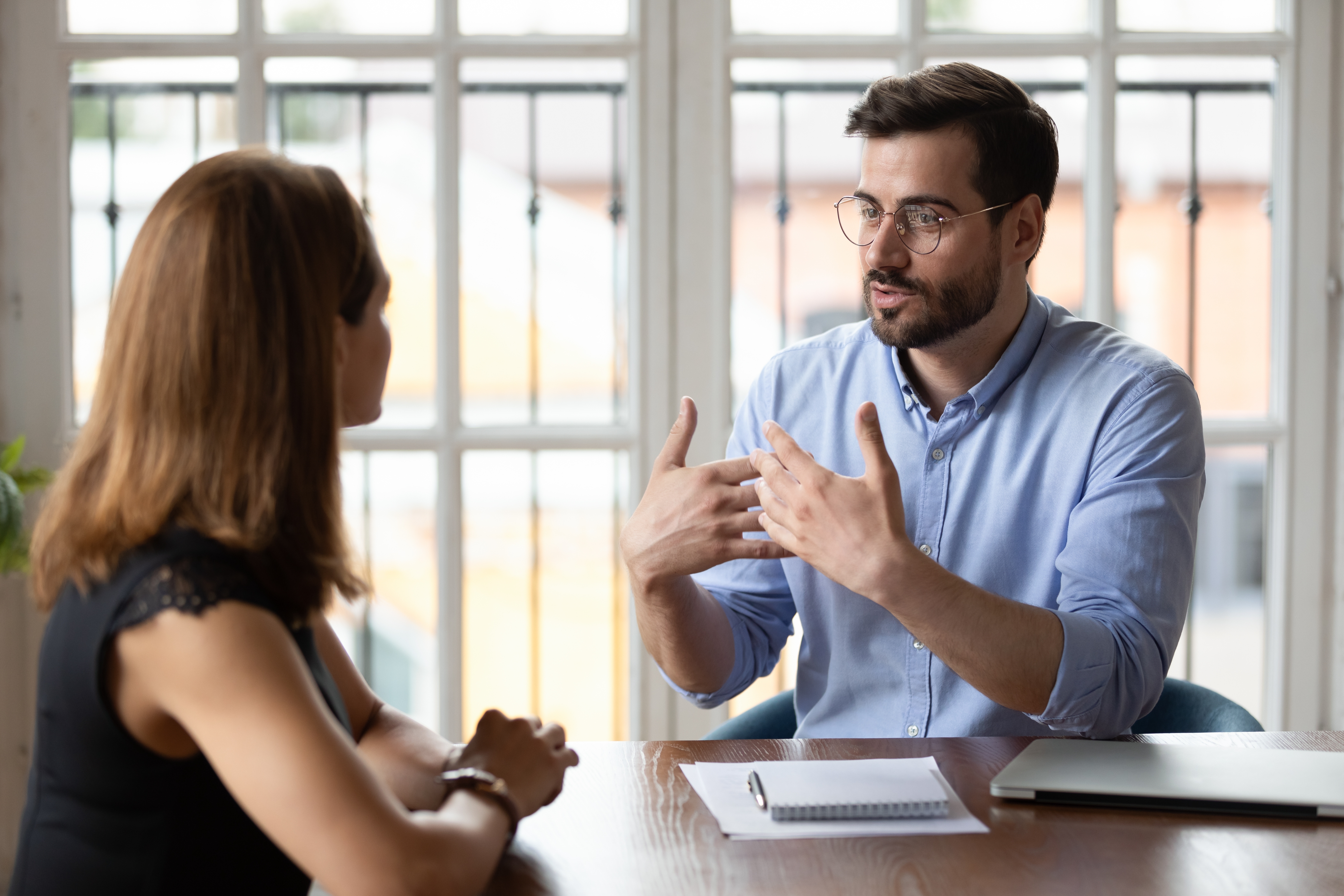 Two people in a discussion at a table, one gesturing while talking. Papers and a laptop are on the table