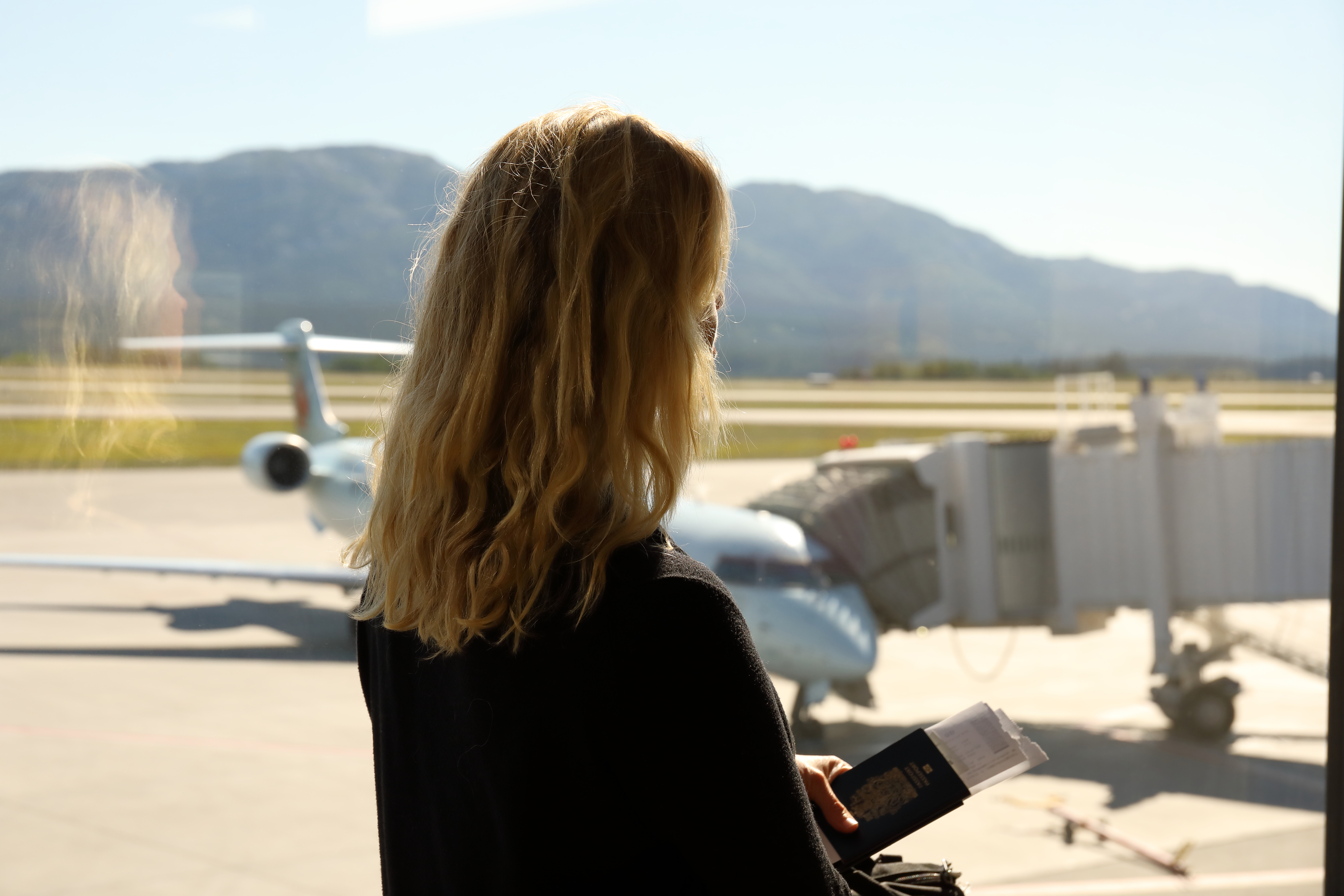 Person with wavy hair stands at airport window, holding a passport, looking at a parked plane on the runway