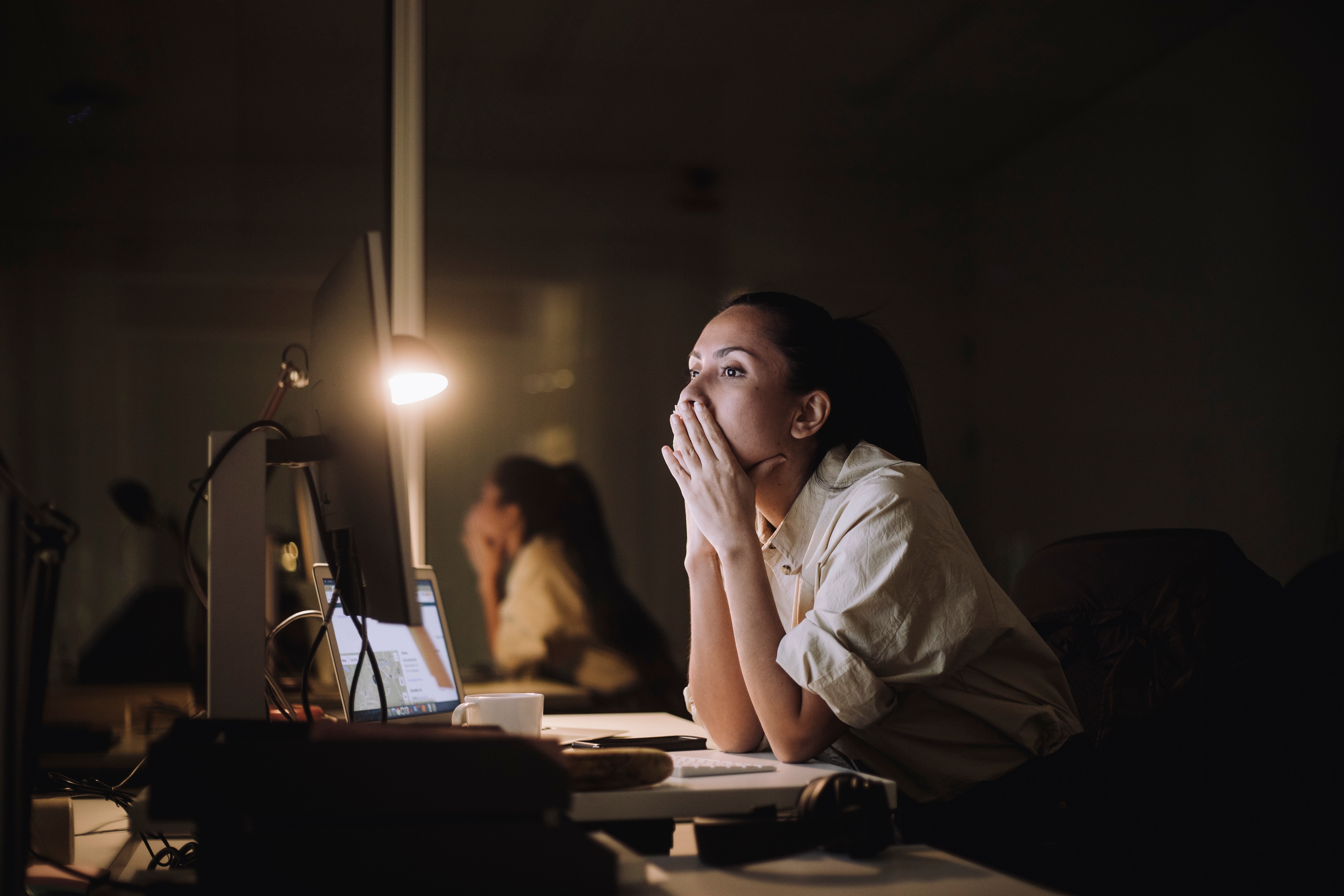 Person in an office works late, staring intently at a computer screen, resting chin on hands. Desk lamp and cluttered workspace visible