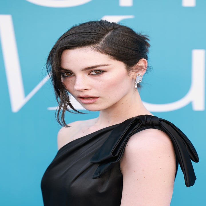 Person in elegant one-shoulder dress with hair styled up, posing in front of a blue backdrop