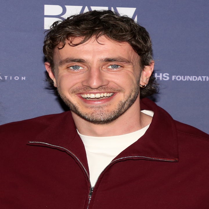 A person smiling at an event, wearing a zip-up jacket over a T-shirt, with a backdrop featuring text and logos