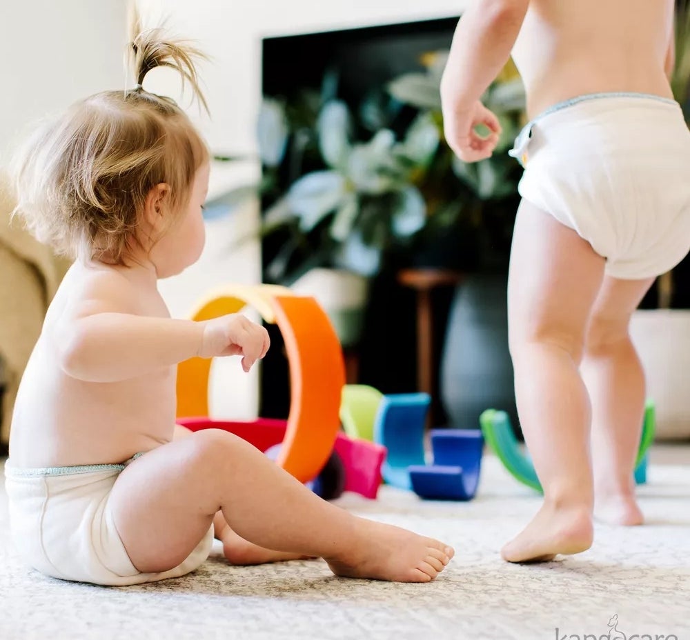 Dua balita yang memakai popok bermain dengan mainan berwarna -warni di lantai ruang tamu