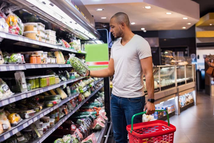 Man holding salad bag in grocery store, wearing a casual T-shirt and jeans, with red basket in hand, standing in front of a produce aisle