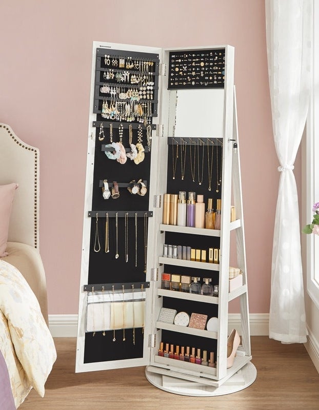 A jewelry cabinet with an open mirrored door displaying earrings, necklaces, and shelves with beauty products in a bedroom setting