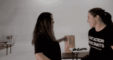 Two women wearing casual black t-shirts share a joyful hug in a minimalistic indoor setting with tables and boxes in the background