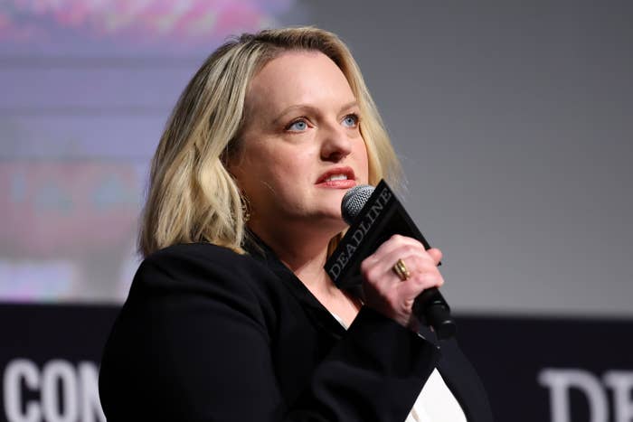 Person speaking on stage, holding a microphone, wearing a black blazer over a white top