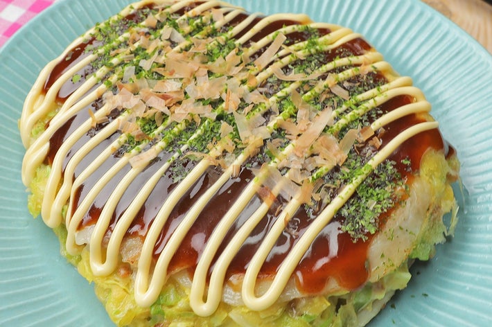 お好み焼きが青い皿に盛られ、マヨネーズとソースがかかっている。周りにはかつお節と青のりが振りかけられている。