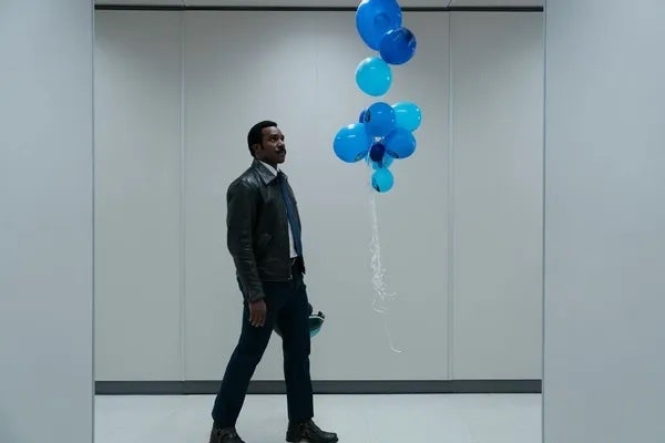 Person in a leather jacket walks in a hallway, looking at floating blue balloons