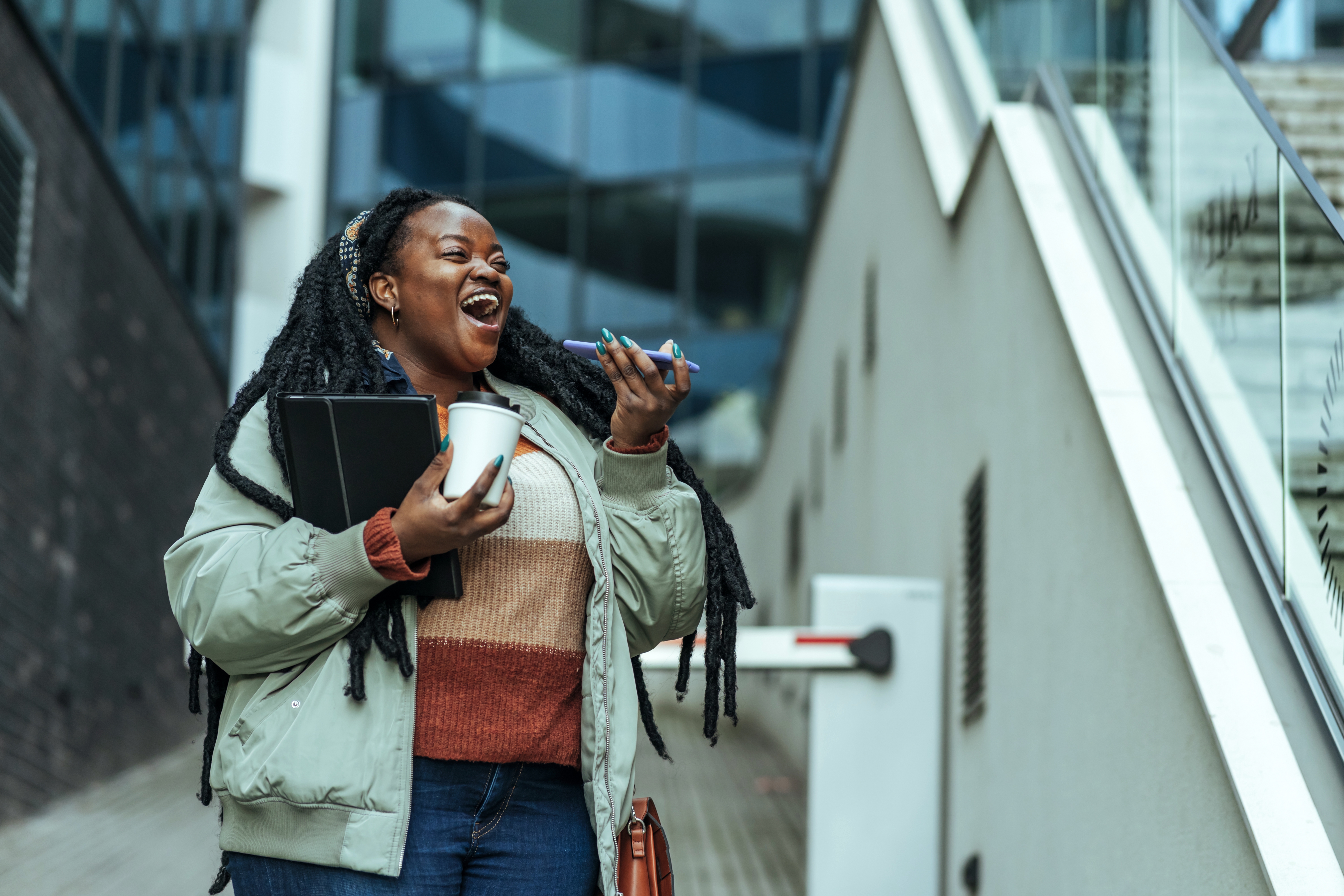 Person smiling and walking outdoors, holding a tablet, coffee cup, and phone near stairs, appearing joyful and engaged in conversation