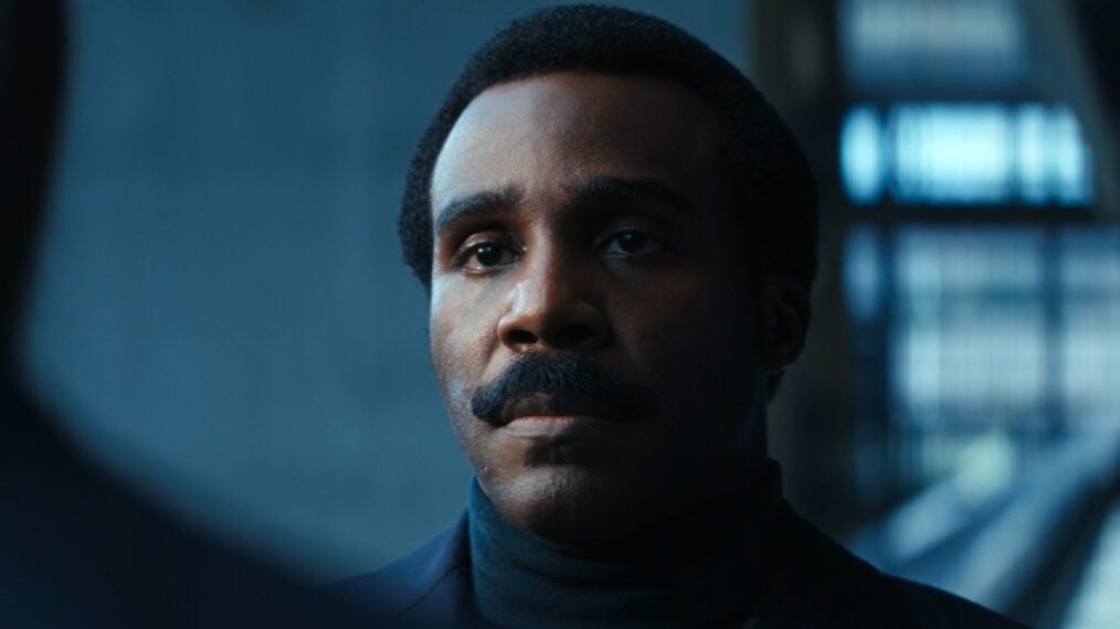 A person with a serious expression, wearing a dark turtleneck and a mustache, in a scene suggesting dramatic tension