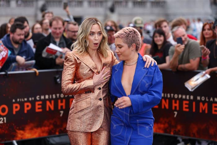 Emily makes a shocked face while using her hand to cover the top of her shiny suit jacket. Florence, who's in a low-cut denim suit jacket and skirt, stands beside her