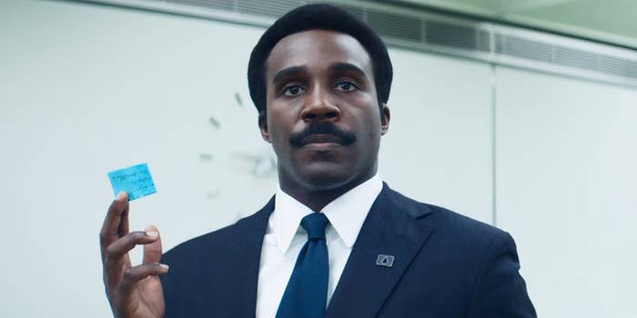 Person in a formal suit holding a small blue note, standing in an office setting with a neutral expression