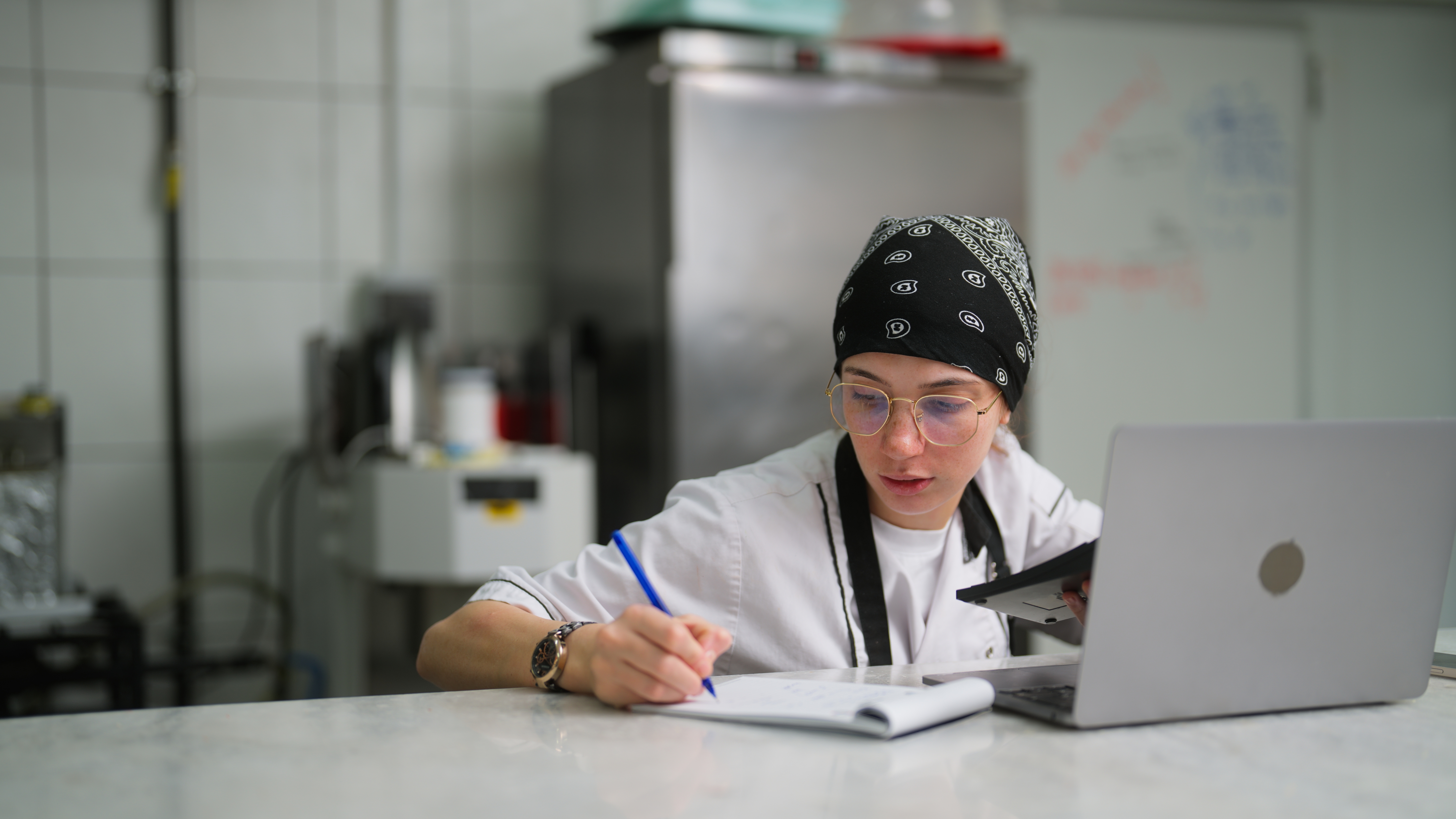 Chef in a kitchen, wearing a bandana and apron, writes on a notepad while looking at a laptop