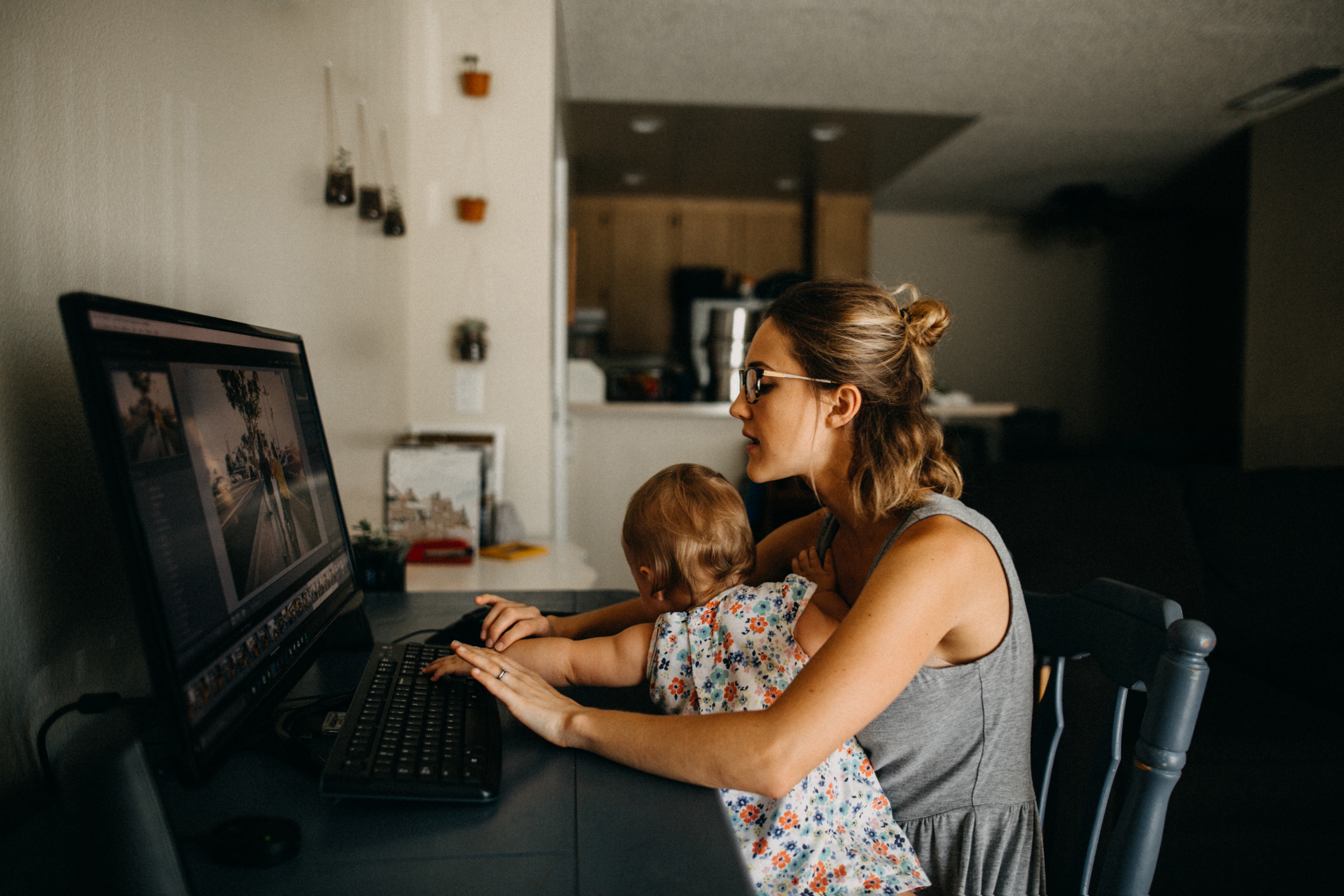Woman multitasks at a computer while holding a toddler. Both focus on the screen in a home setting, suggesting a blend of work and caregiving