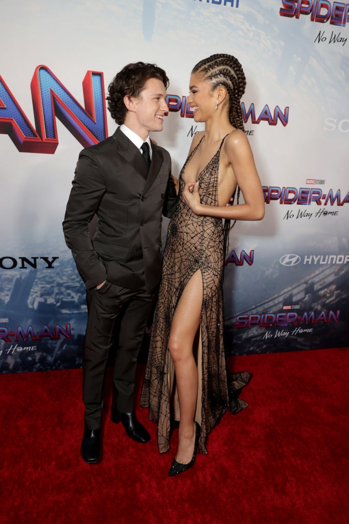 Tom in a suit and Zendaya in a spider web dress, facing each other on the red carpet