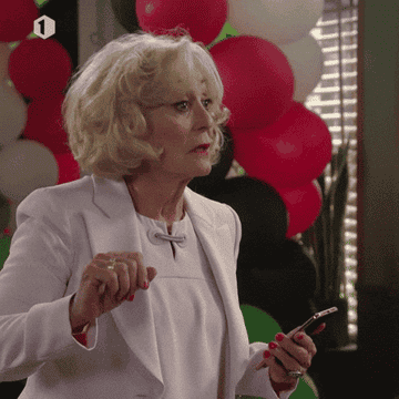 Woman looking surprised at a party, holding a smartphone. Balloons are visible in the background