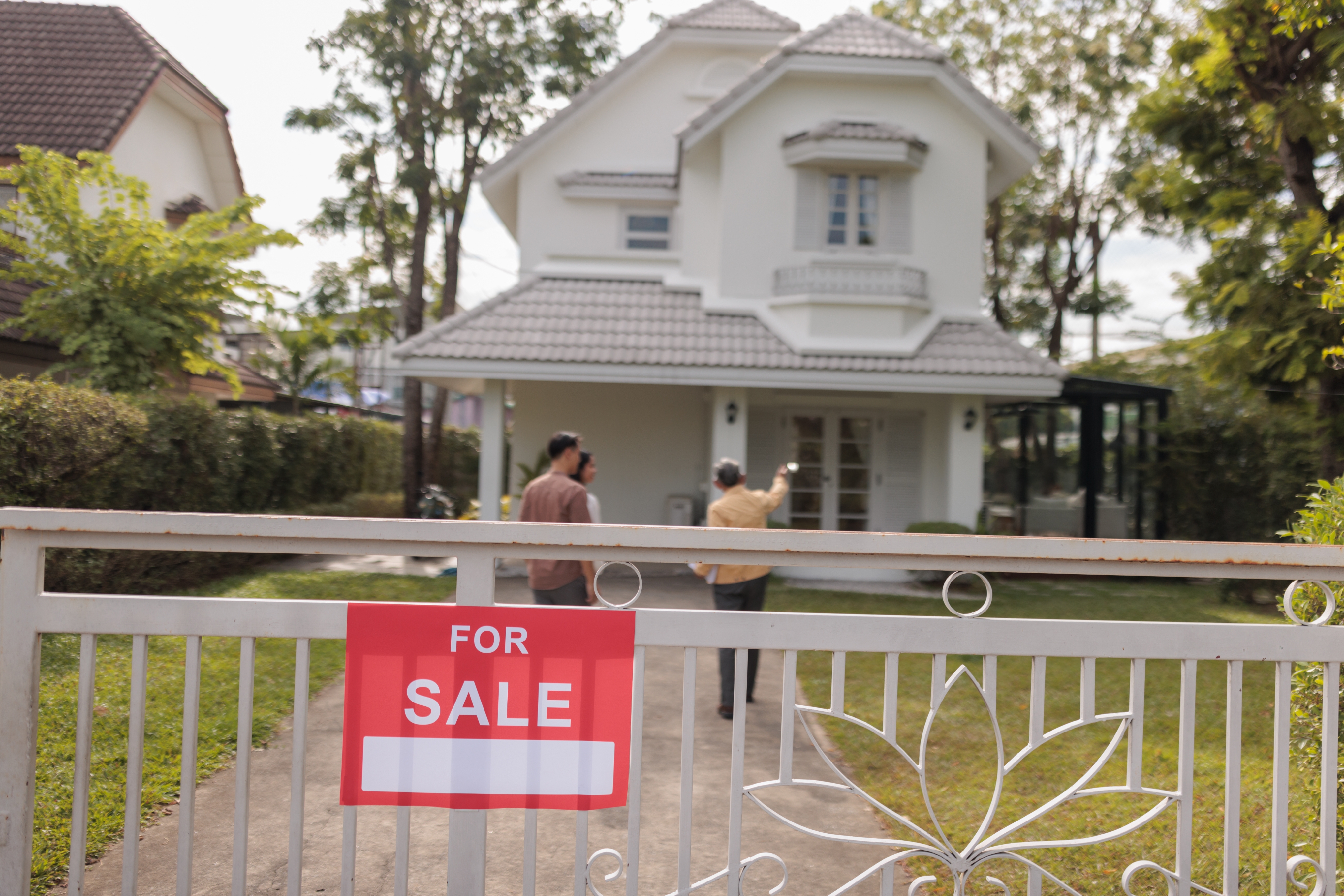 Two people stand near a 'For Sale' sign in front of a large house with a garden