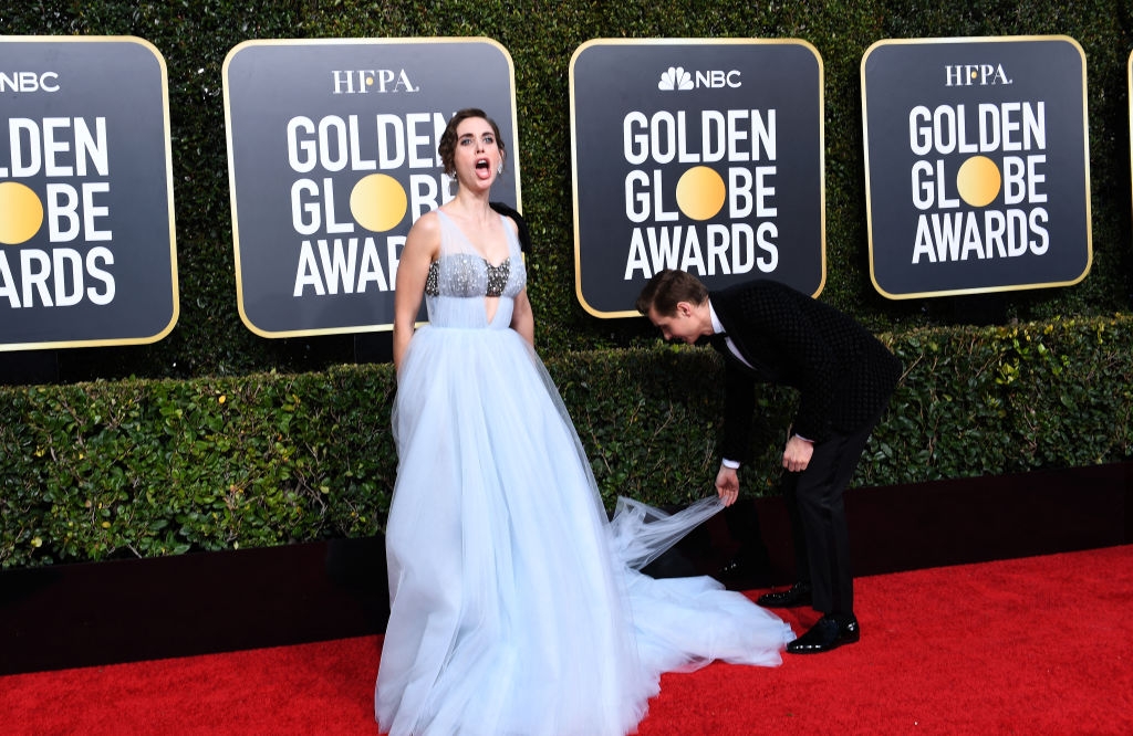 Alison in an elegant tulle gown on a red carpet, with Dave adjusting the dress train, which is caught on a hedge