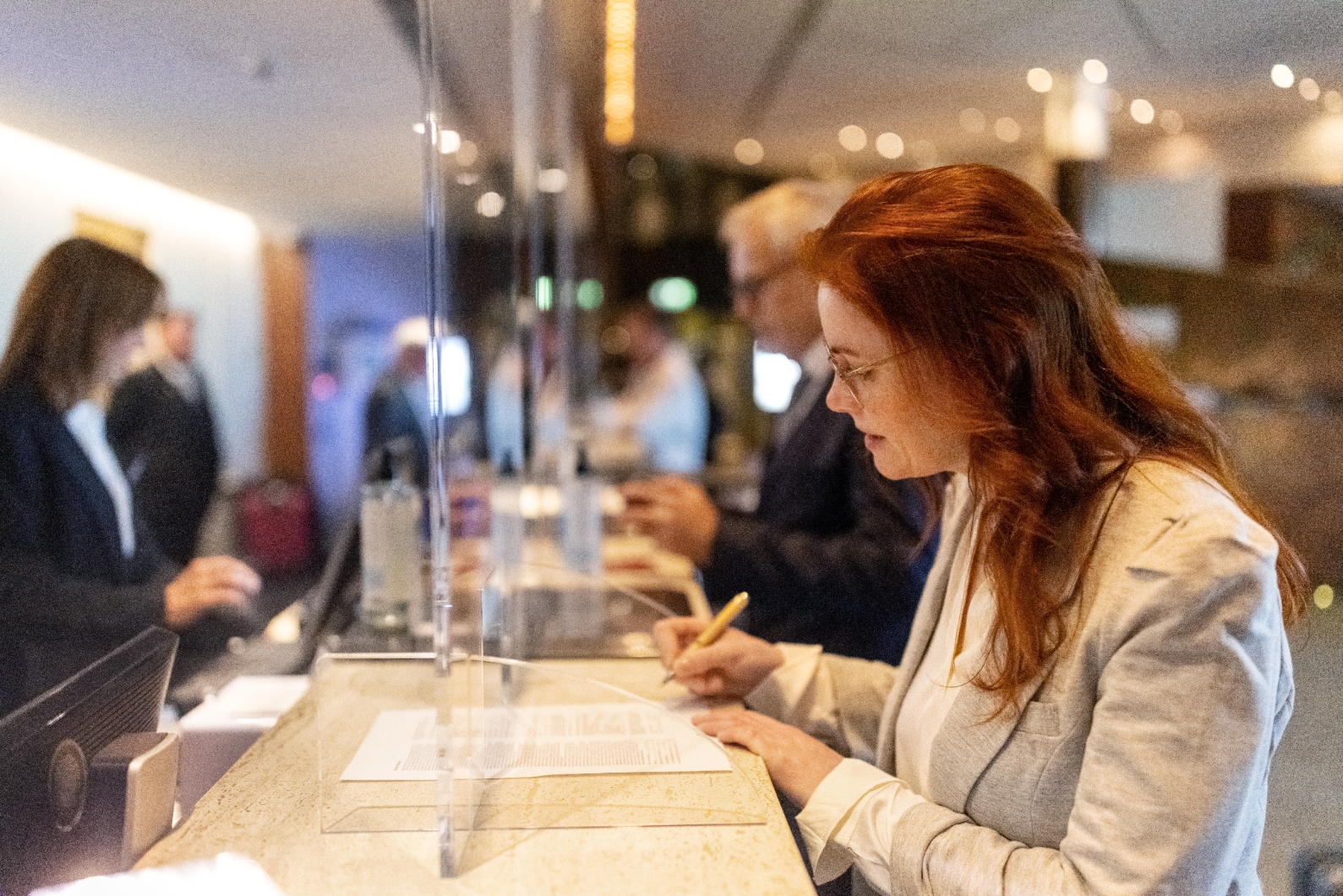 A person in a suit fills out paperwork at a hotel reception desk, with staff assisting other guests in the background