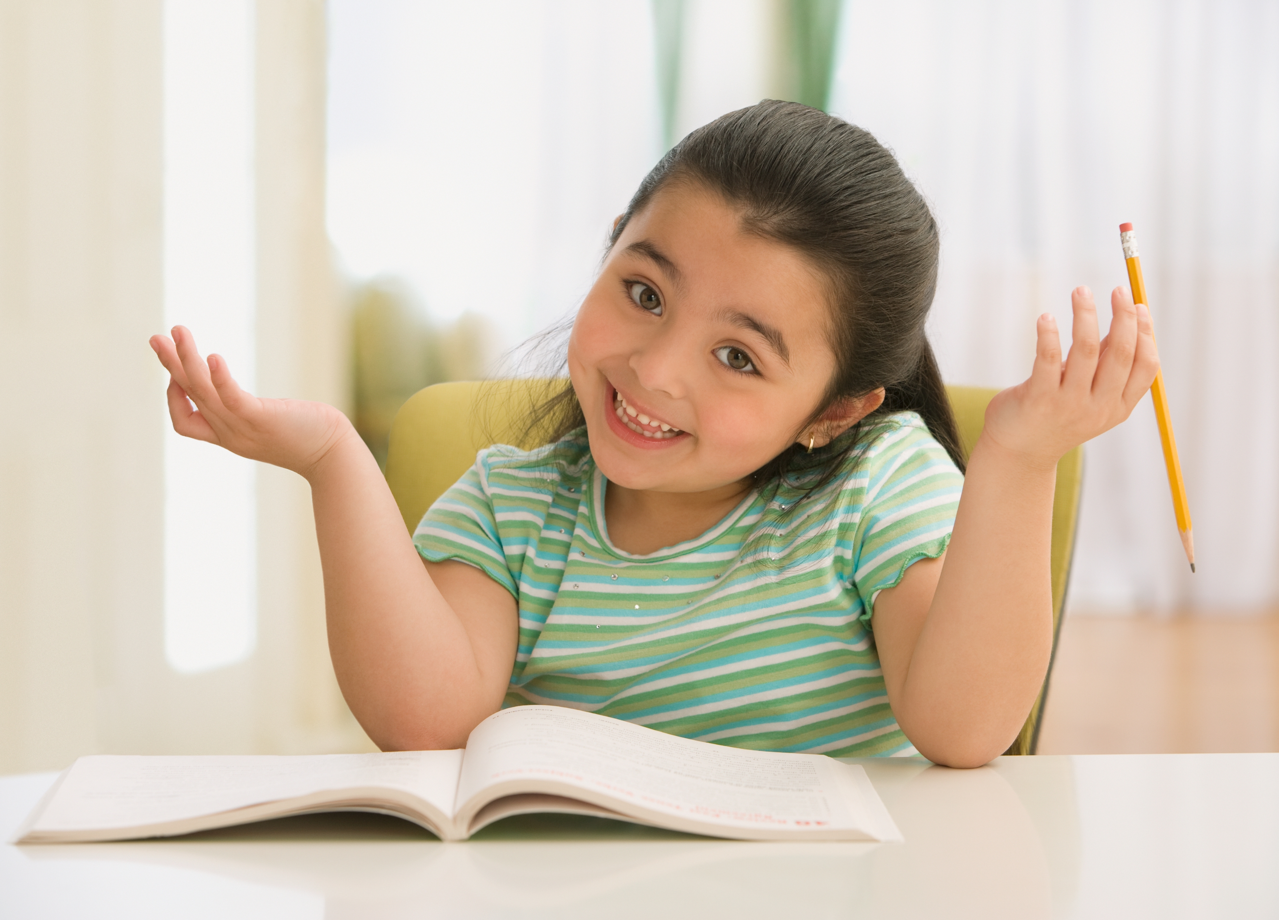Child shrugging with open book and pencil at a table, smiling with a playful expression