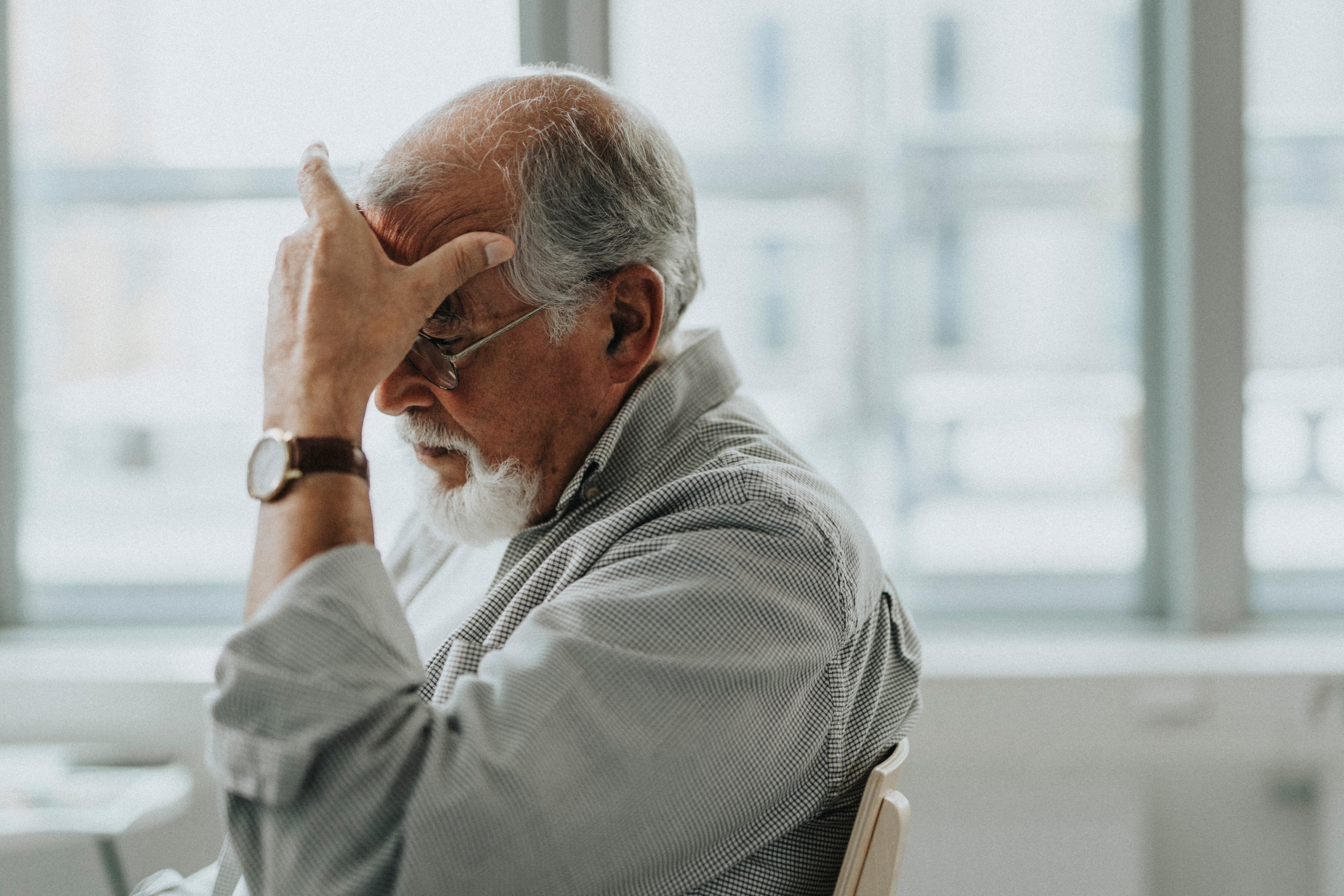 Older man with glasses, sitting in thought, hand on forehead. He wears a watch and a striped shirt. Background shows blurred windows