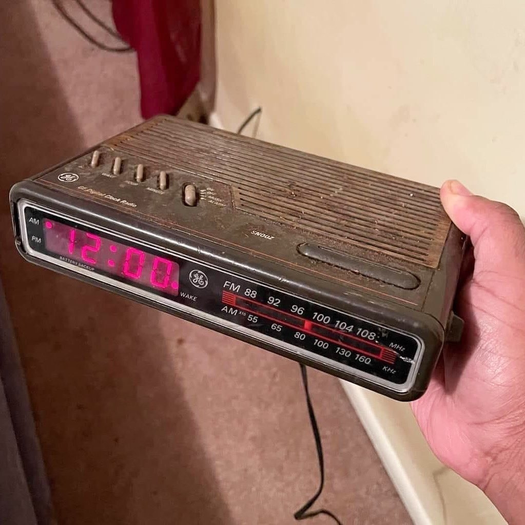 A person holds a vintage digital clock displaying 12:00, featuring dials for FM and AM radio frequencies