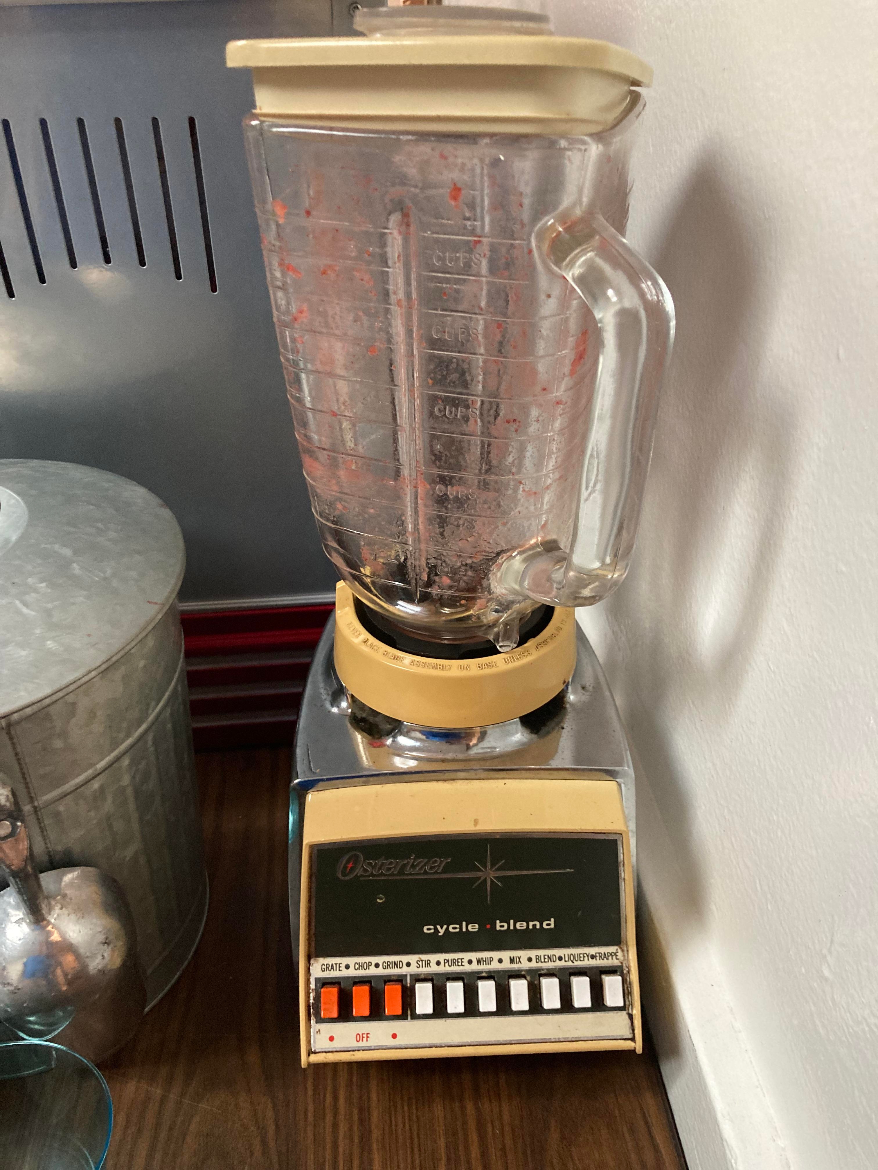 Vintage Osterizer blender with glass jar, pink residue inside, and retro control panel on a countertop next to a metal container
