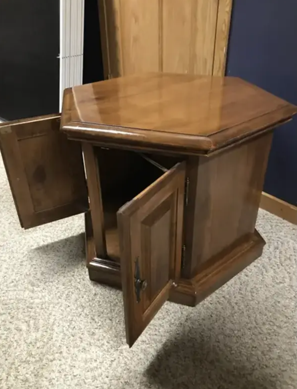 Hexagonal wooden side table with open cabinet doors, revealing empty storage space inside. Carpeted floor visible