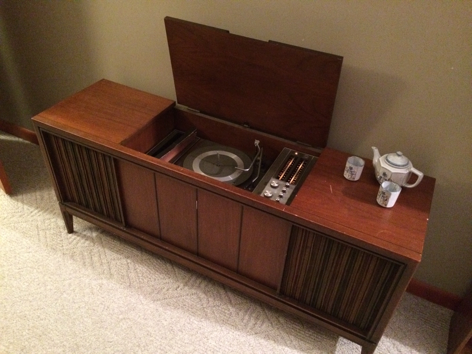 A vintage wooden record player cabinet with an open lid, revealing a turntable and control panel. A teapot and two cups sit on top