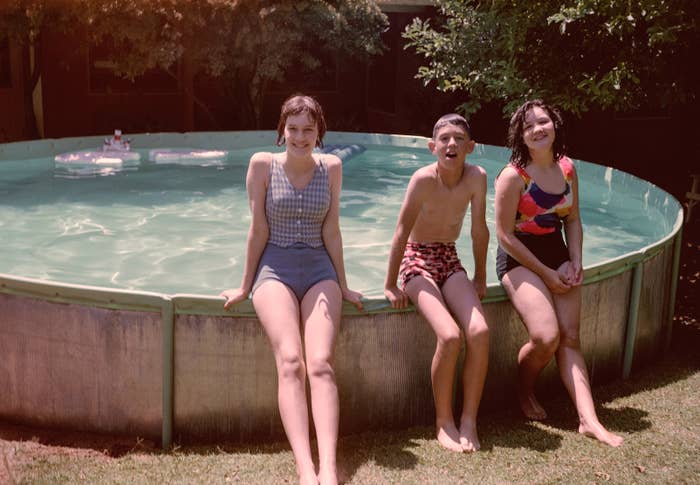 Three people, two in swimsuits and one in swim trunks, sit on the edge of a circular above-ground pool, smiling and enjoying a sunny day