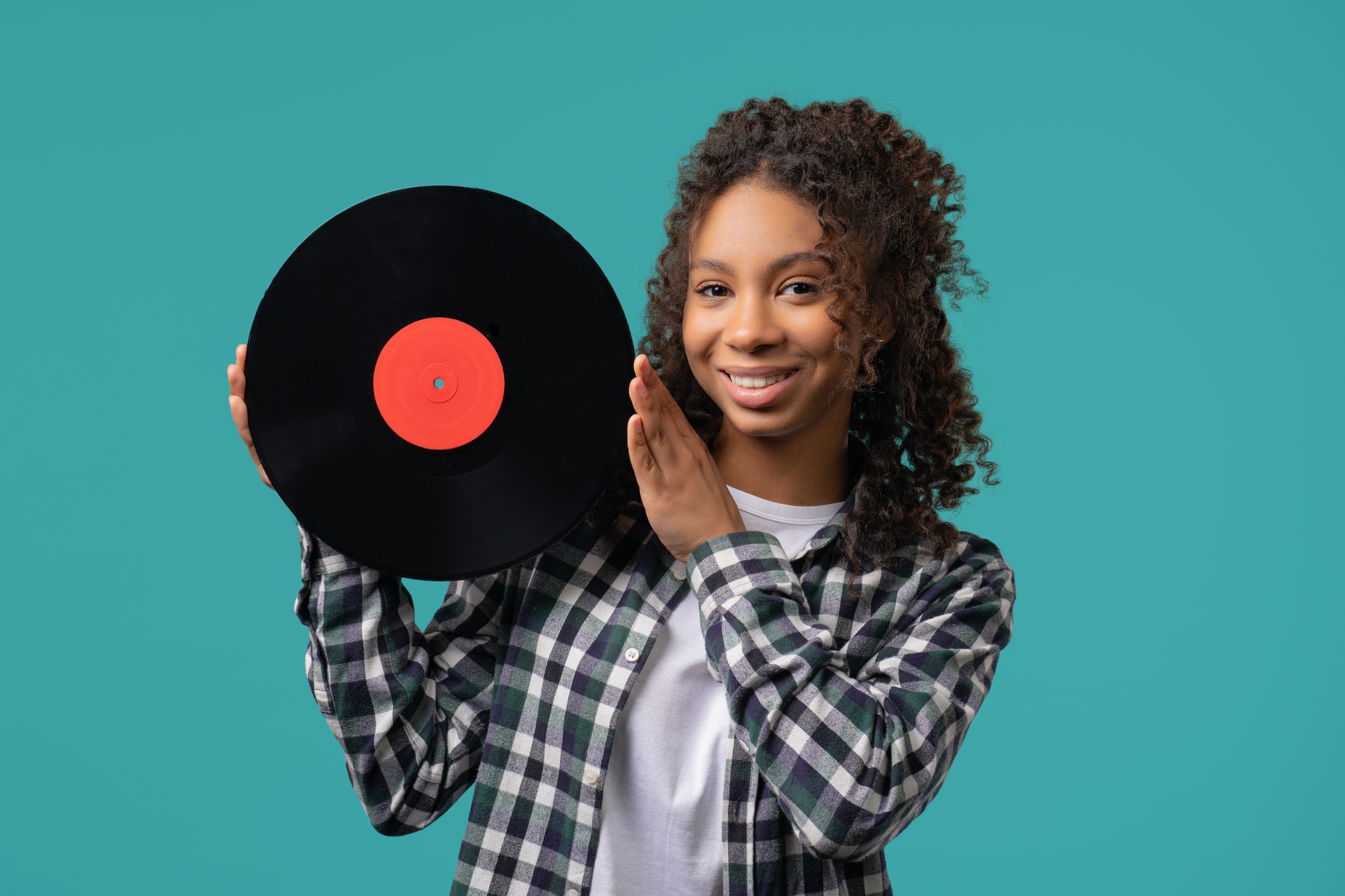 Person smiling, holding a vinyl record in one hand, dressed in a casual flannel shirt over a plain tee