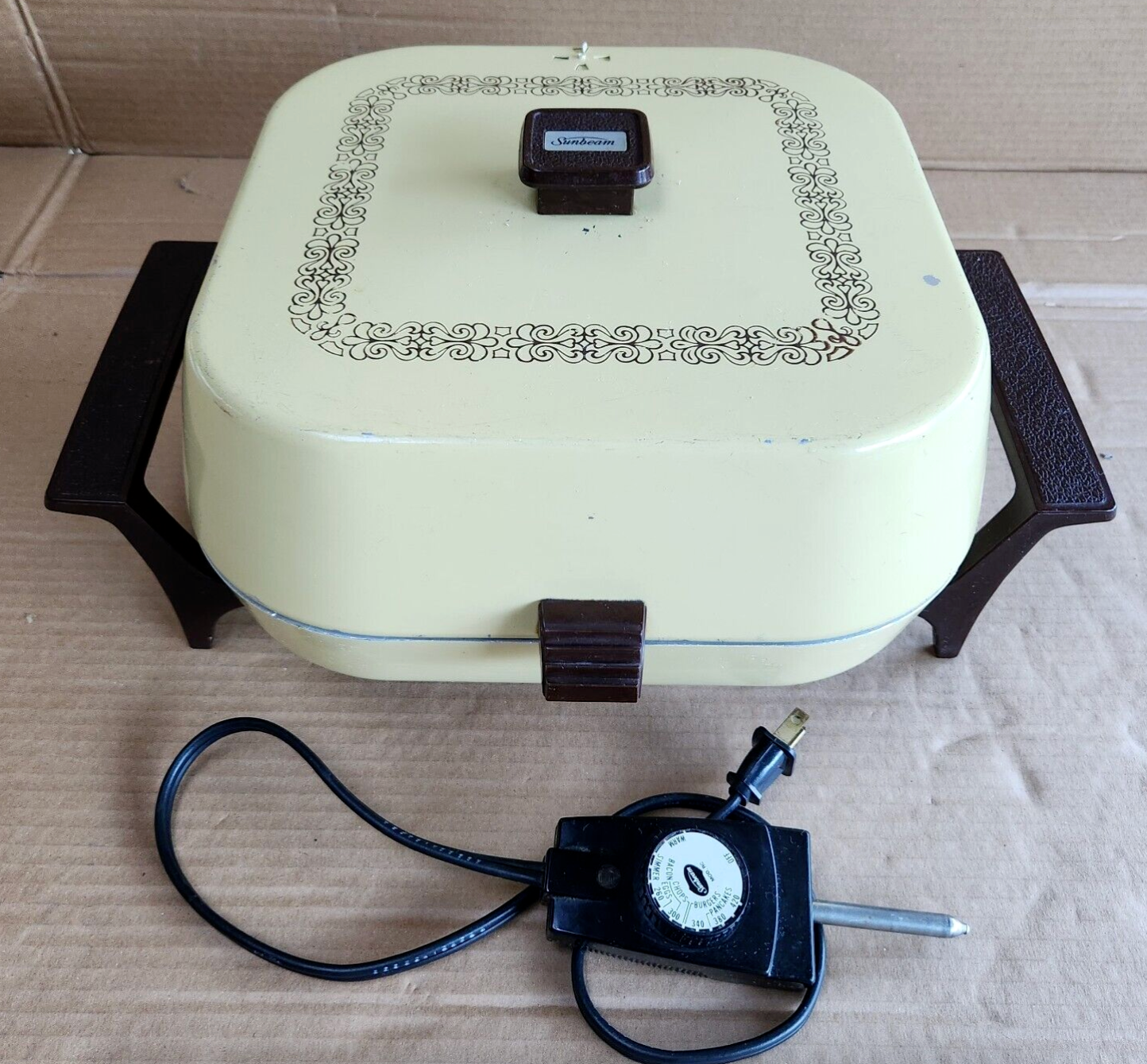 A vintage electric skillet with handles, ornate border design on the lid, and an attached temperature control knob on a cardboard surface