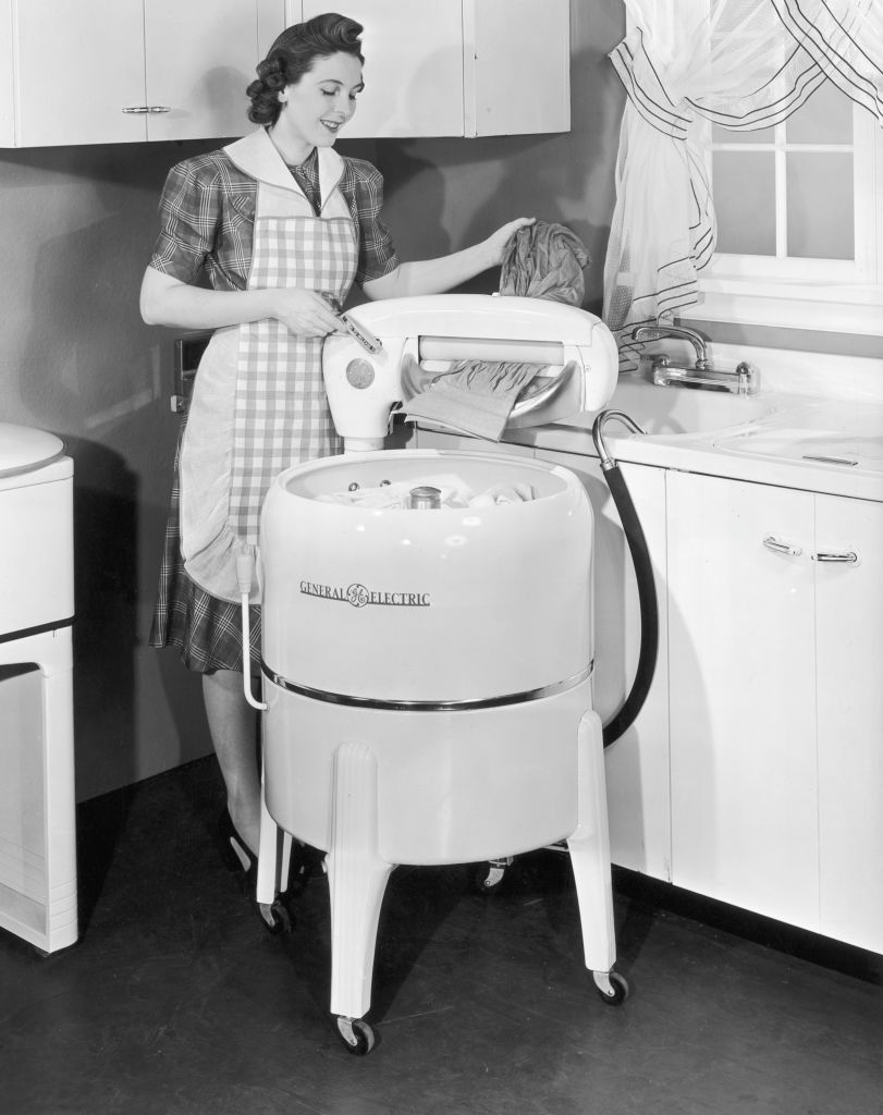 A 1950s woman operates a vintage General Electric washing machine, wearing a checkered dress and apron in a domestic kitchen setting