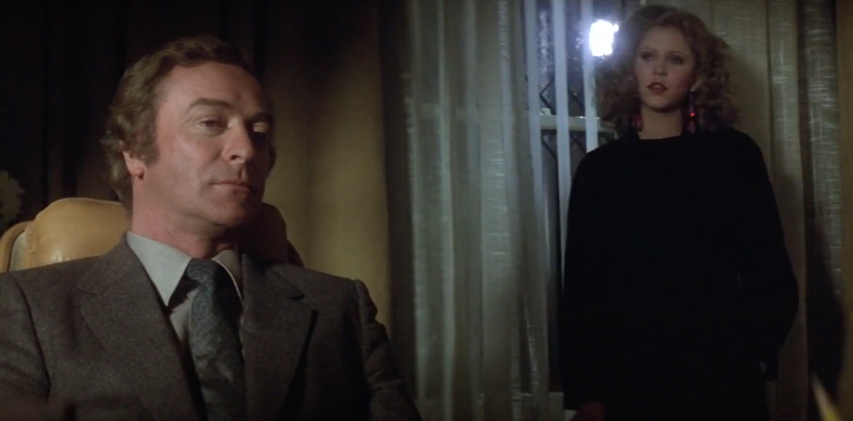 A man in a suit sits pensively in a dim room, while a woman with curly hair stands nearby, wearing a dark outfit and earrings