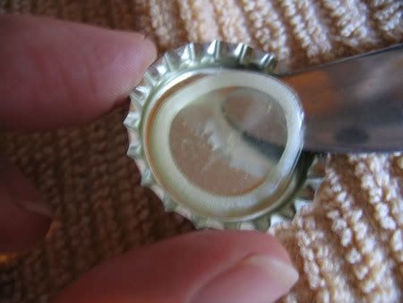 A bottle cap being lifted with a spoon, showing the inside seal. Fingers hold the cap on woven fabric