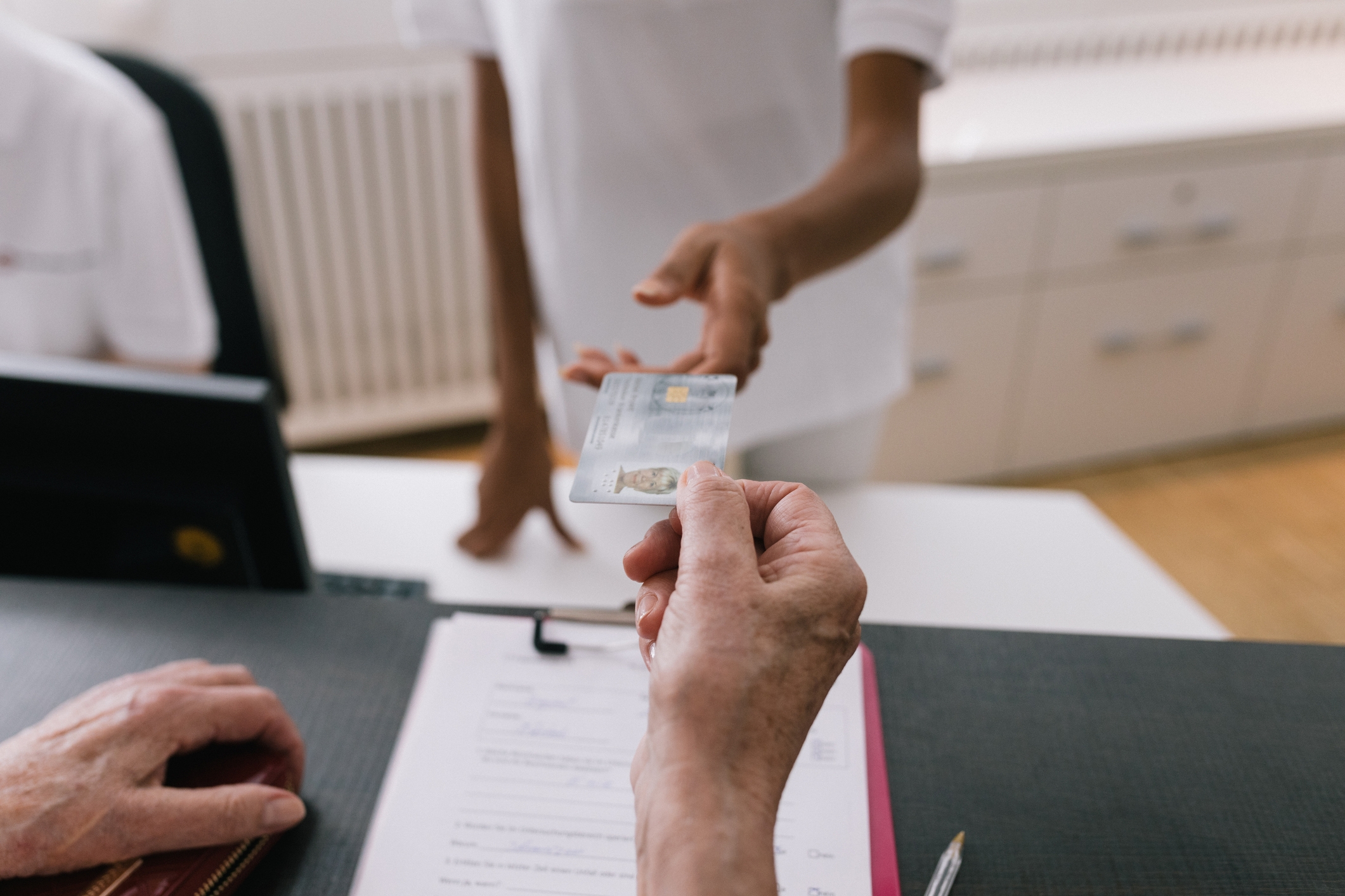 Person som overleverer et forsikringskort til en annen person på tvers av et skrivebord med dokumenter, noe som antyder en transaksjon eller innsjekkingsprosess
