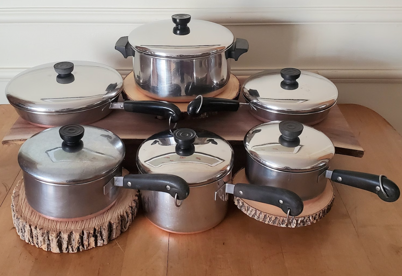 A set of five stainless steel pots and pans with black handles, arranged on a dining table with wooden trivets underneath