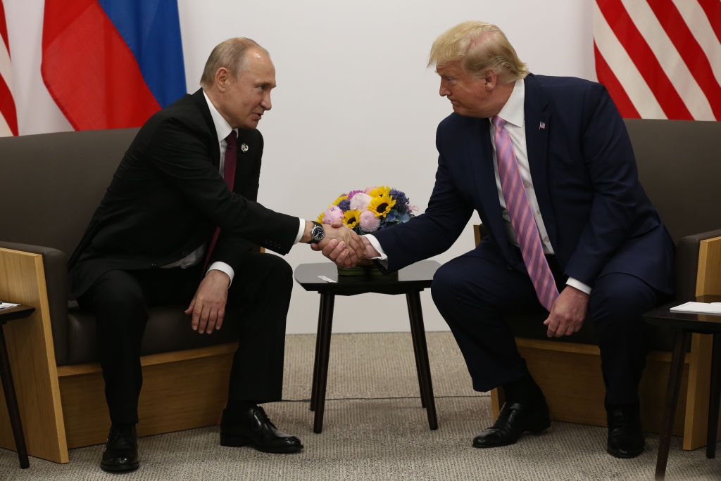 Two men in suits sit and shake hands during a formal meeting, with flags and flowers in the background