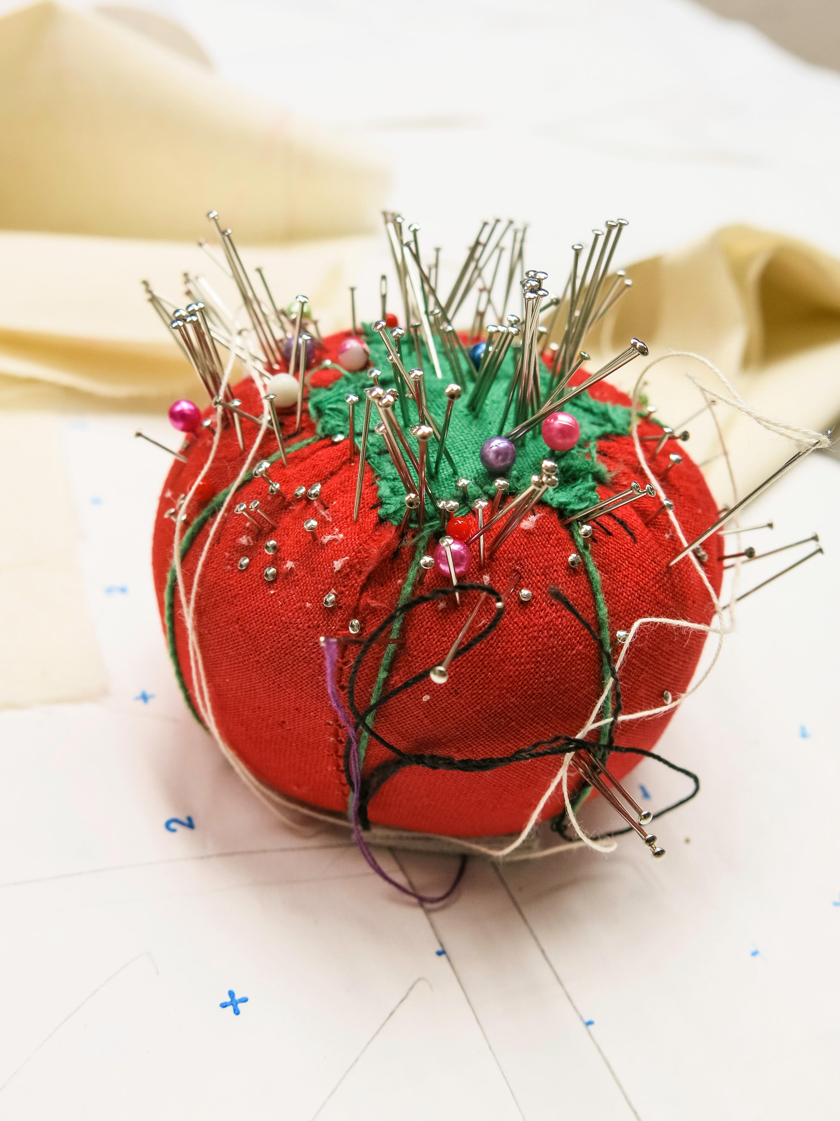 Pin cushion shaped like a tomato, filled with assorted sewing pins and needles, surrounded by threads