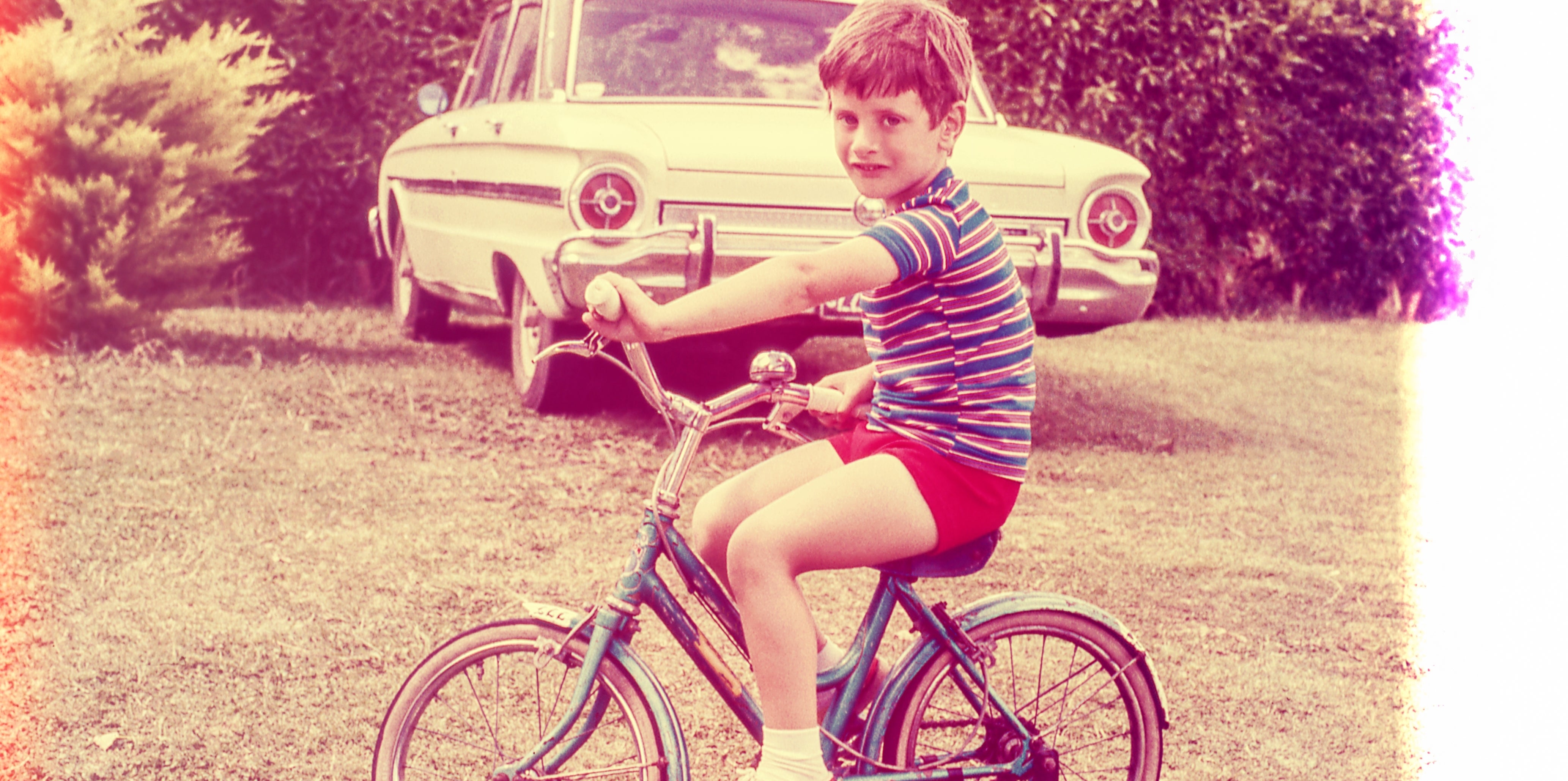 Child on a bicycle in a grassy yard with an old car in the background. Photo has a vintage effect