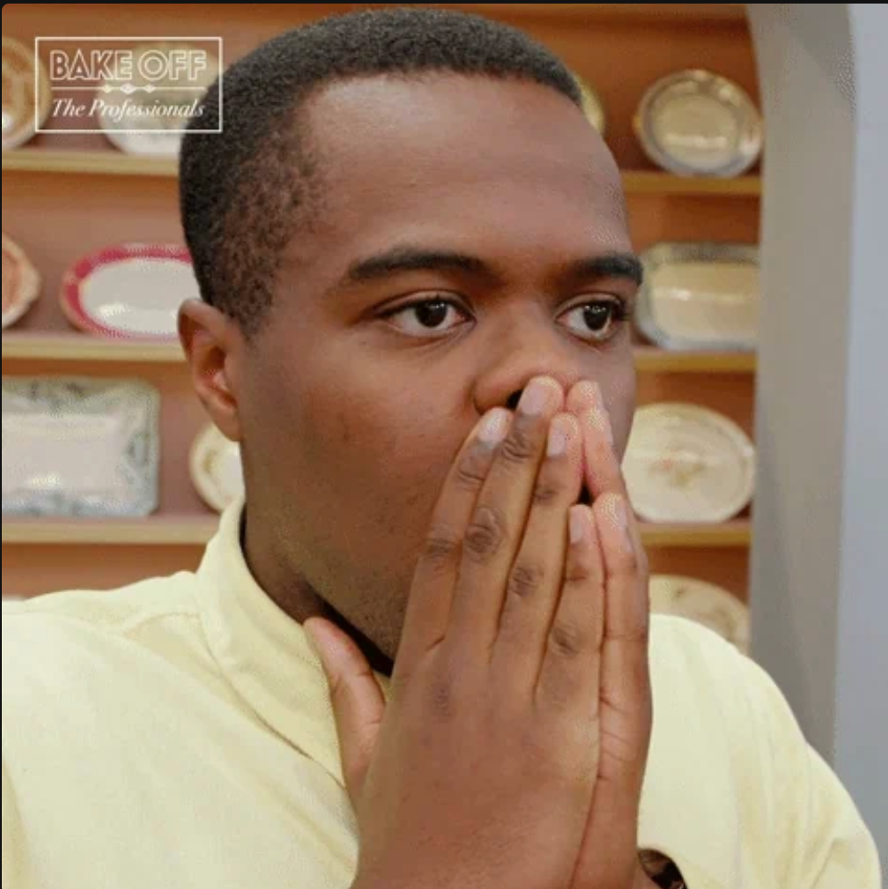 A person, appearing surprised with hands over mouth, stands in a kitchen-themed setting with decorative plates in the background