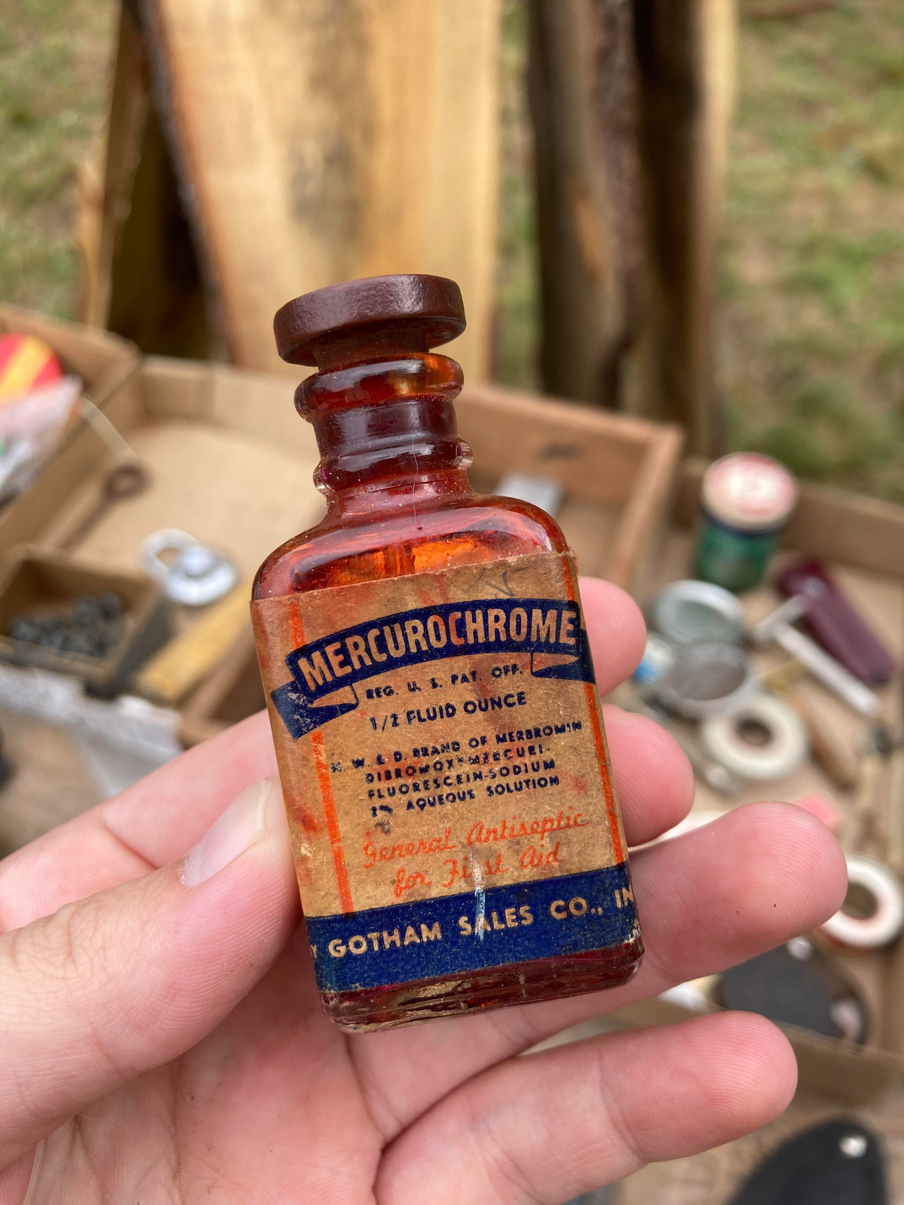 A vintage bottle of Mercurochrome with label details held in a person's hand, with assorted tools and items visible in the background
