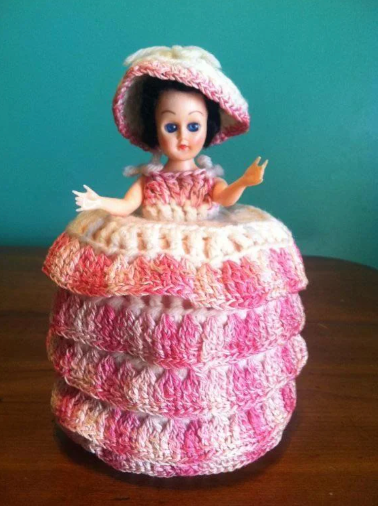 Doll with a crocheted dress and bonnet, layered skirt, arms raised. It's placed on a wooden surface against a plain backdrop