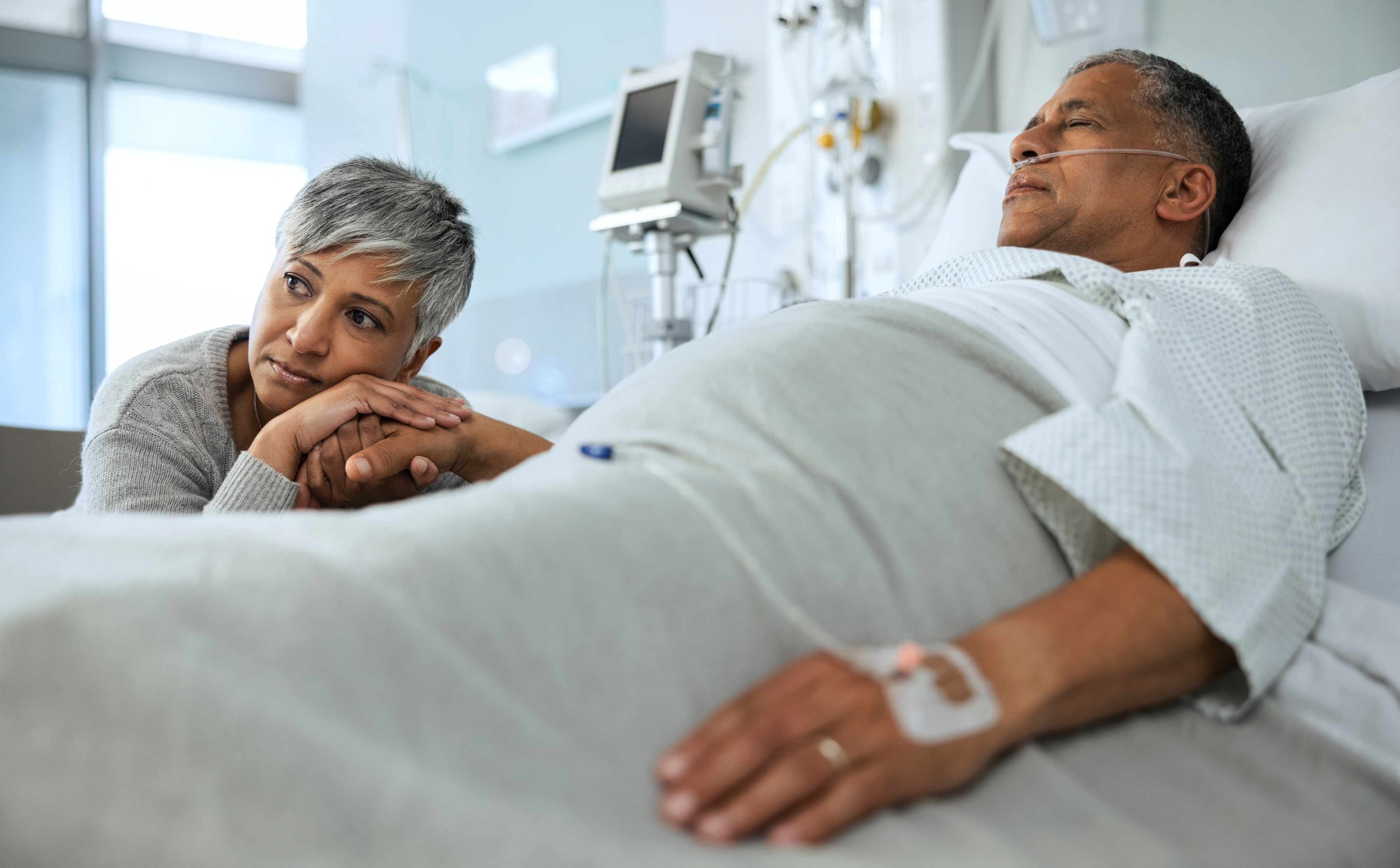A woman holds the hand of a man lying in a hospital bed, both appearing calm and contemplative. Medical equipment is visible in the background