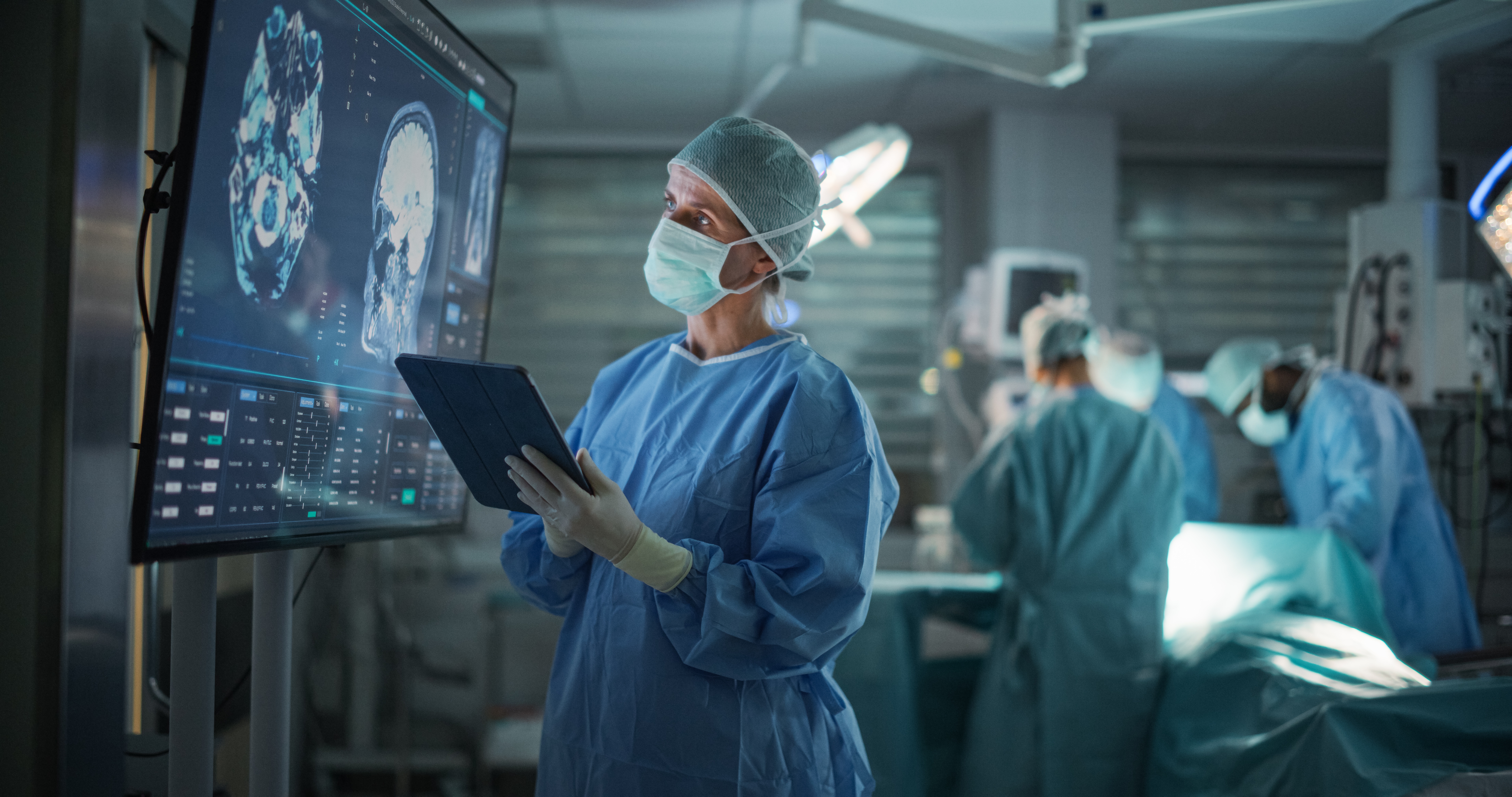 Doctor in surgical attire reviews scans on a screen in an operating room while a surgical team works nearby