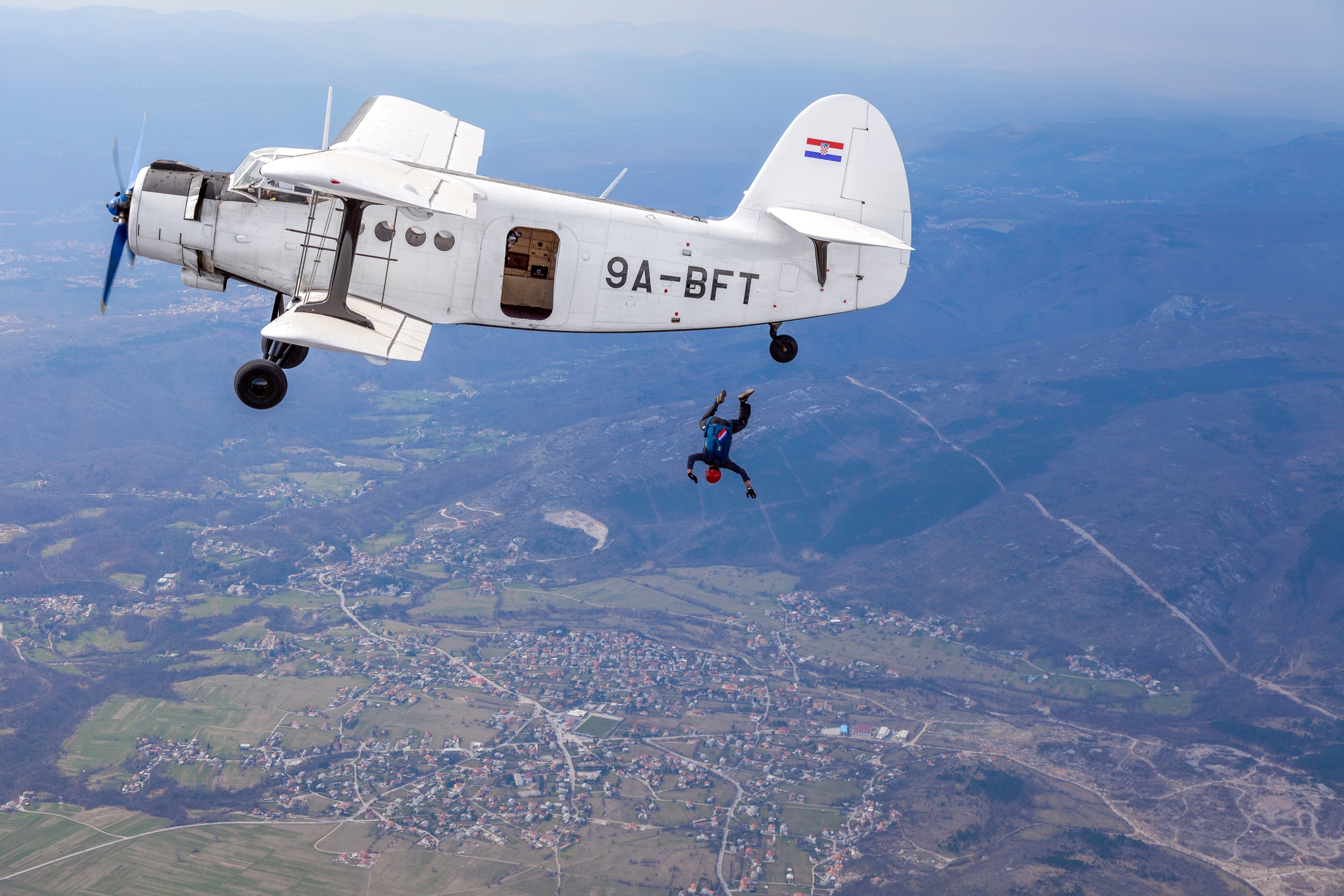 Skydiver jumps from a small propeller plane over a cityscape, mid-air above a sprawling landscape