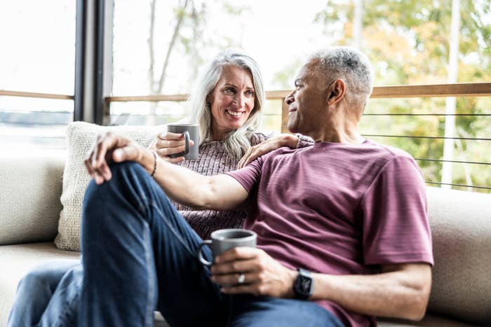 Older couple sitting on a sofa, smiling at each other while holding coffee mugs, conveying warmth and connection
